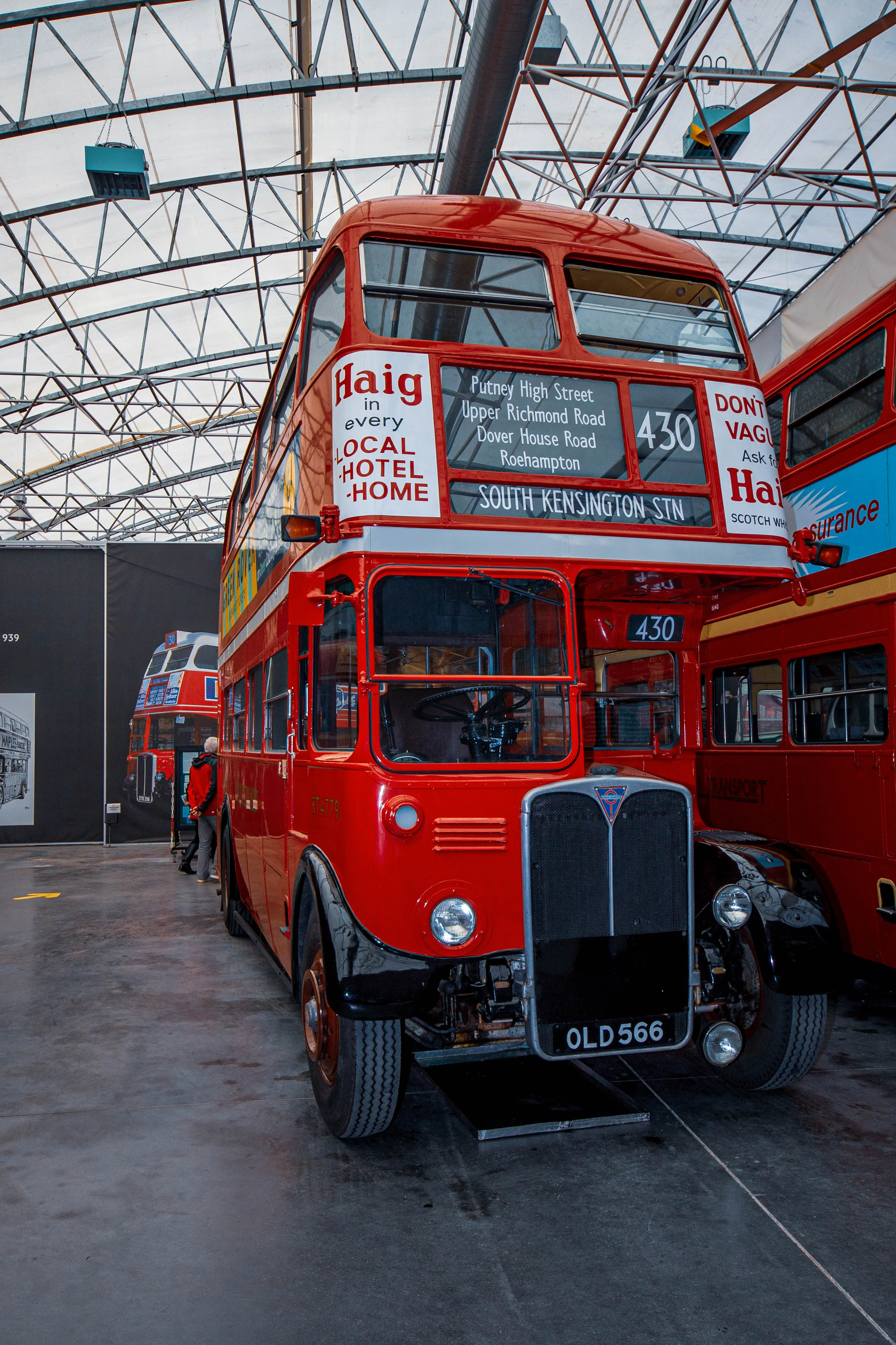 A bright red, vintage double-decker bus with black trim is parked indoors. The upper deck displays route information for South Kensington Station and adverts for 