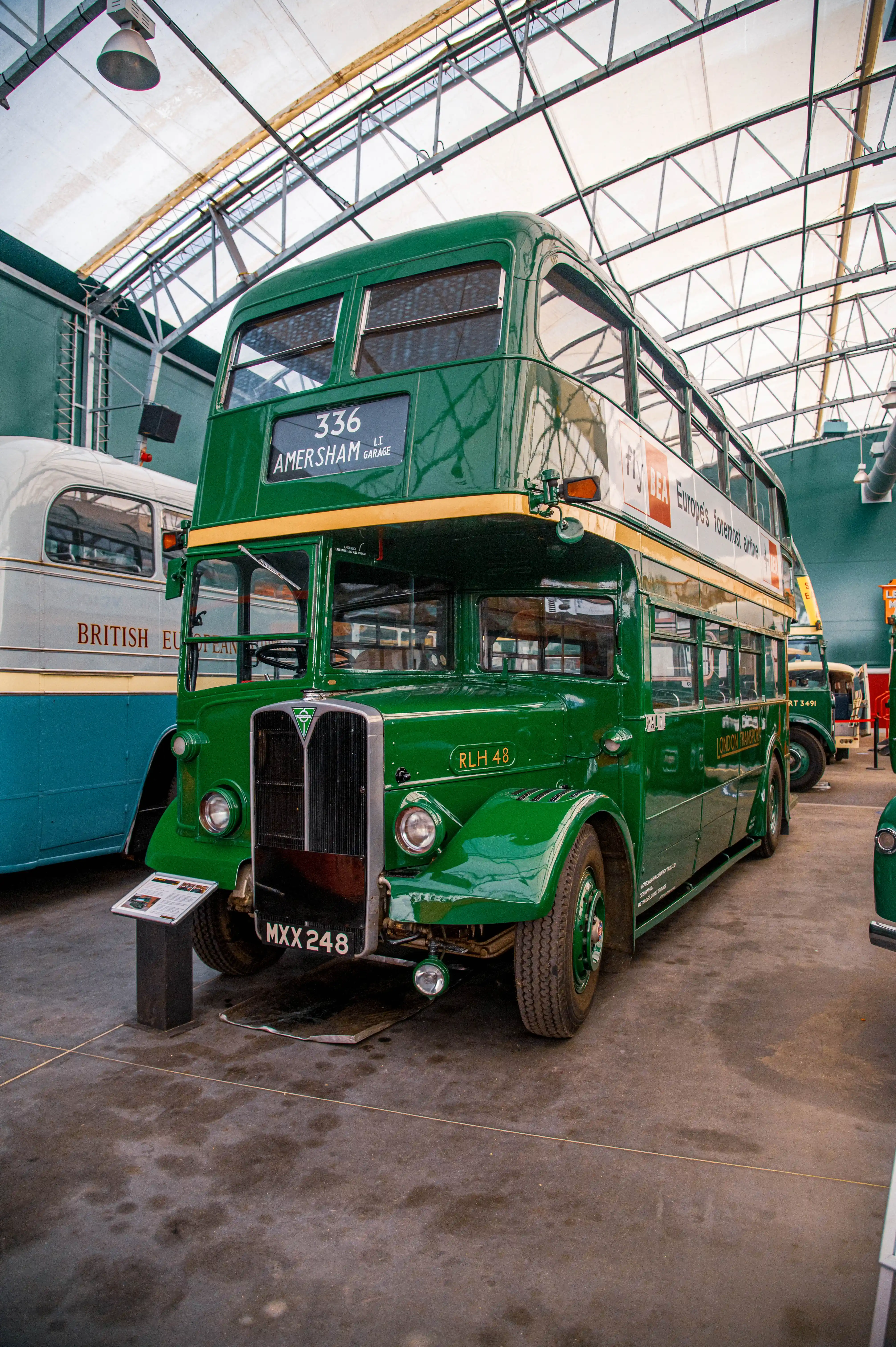 A bright green, vintage London double-decker bus with a yellow stripe is parked inside a building. The bus has the route number 336 and destination Amersham LT Garage displayed at the front. Another bus with BRITISH EUROPEAN AIRWAYS visible is to its left.