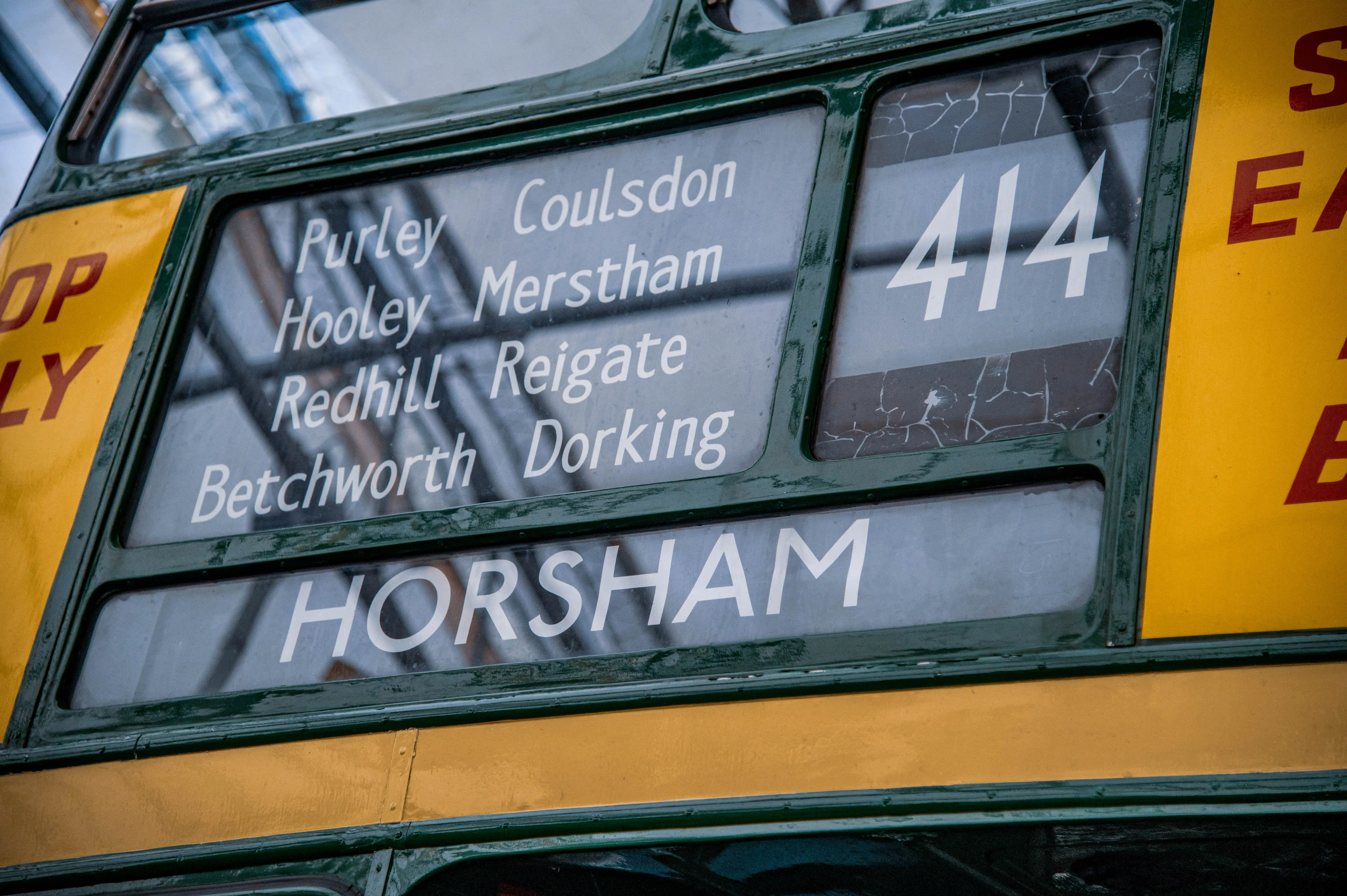 Close-up of a dark green and yellow vintage bus exterior showing a destination board with white text. The board lists multiple locations: Purley, Coulsdon, Hooley, Merstham, Redhill, Reigate, Betchworth, and Horsham. The route number 414 is visible in a panel with cracked glass. Yellow panels with red lettering reading STOP ONLY and EAST are partially visible.