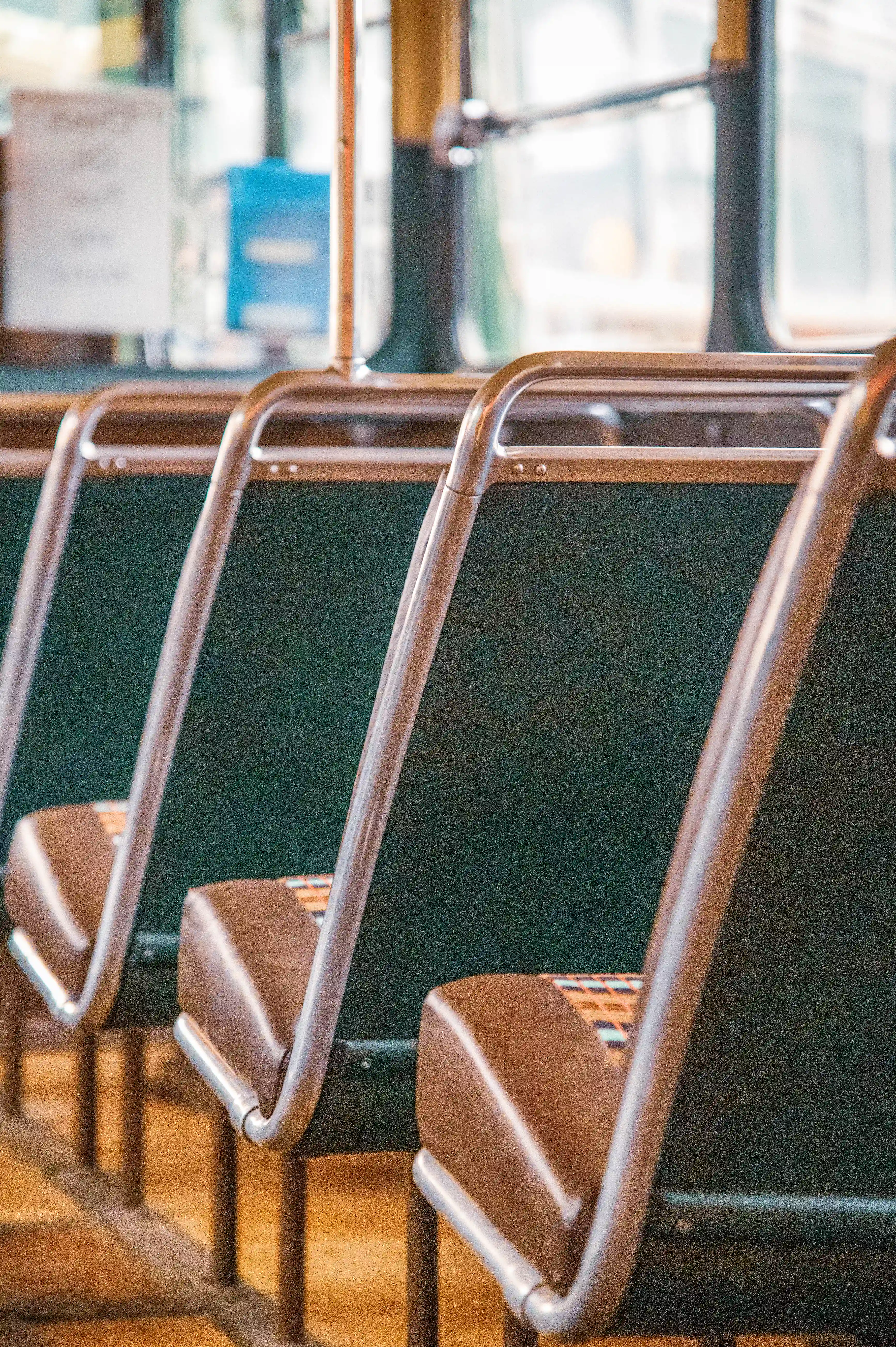 Close-up view of unoccupied bus seats arranged in a row. The seats have dark green fabric backs and brown padded headrests, with shiny metal frames.