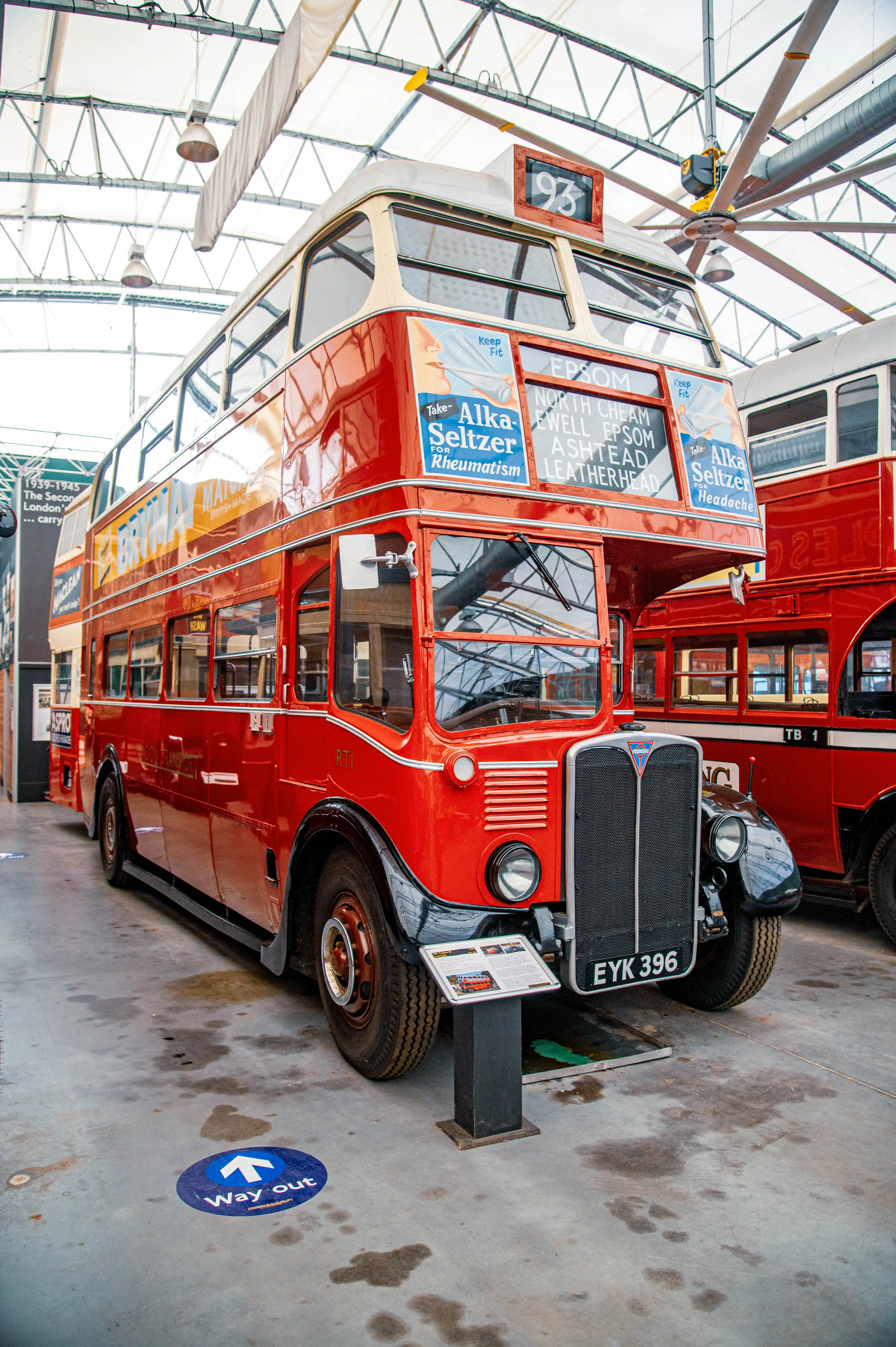 Front three-quarter view of a bright red, vintage double-decker bus with cream trim, parked indoors. Advertisements for Alka Seltzer are visible on its side, and a route display lists destinations. A 