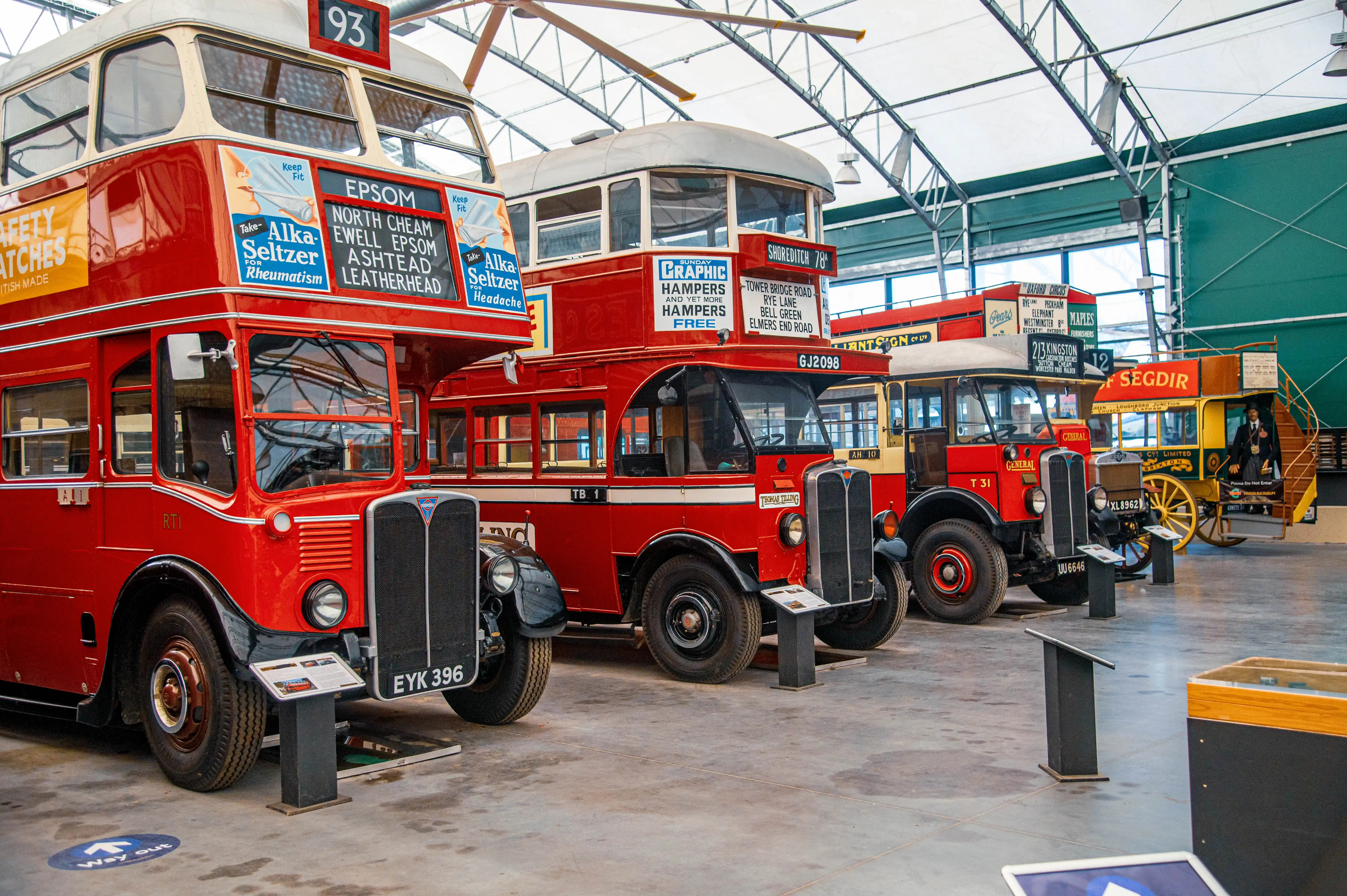 A row of vintage red double-decker buses and a yellow horse-drawn carriage are parked inside a museum or exhibition hall.