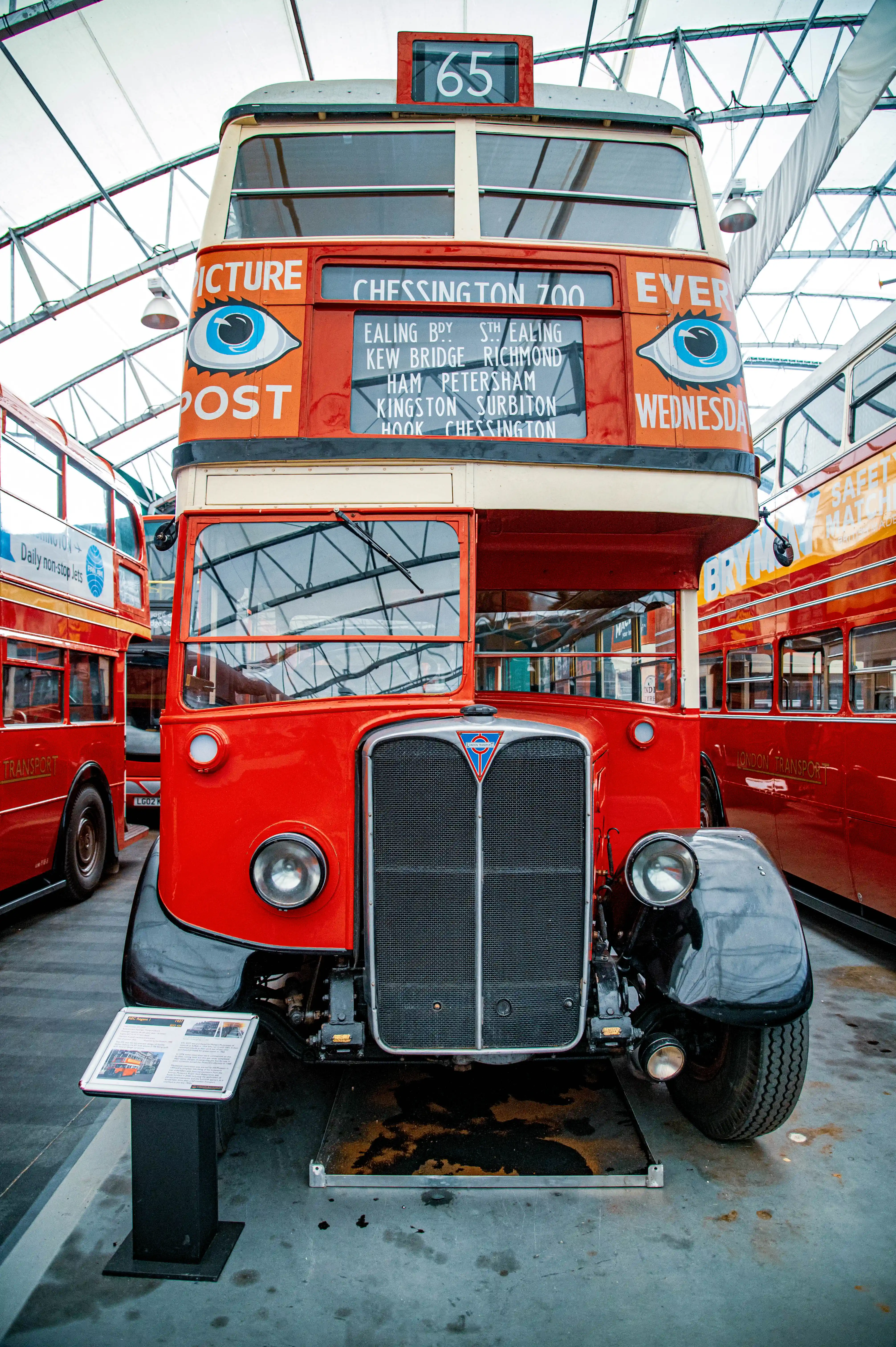 Front view of a bright red vintage AEC Routemaster double-decker bus with cream trim. It displays the route number 65 and a destination board listing various London locations such as Ealing, Richmond, and Kingston. The bus has large circular headlights and a prominent grille. A sign with text and images is placed in front of the bus.