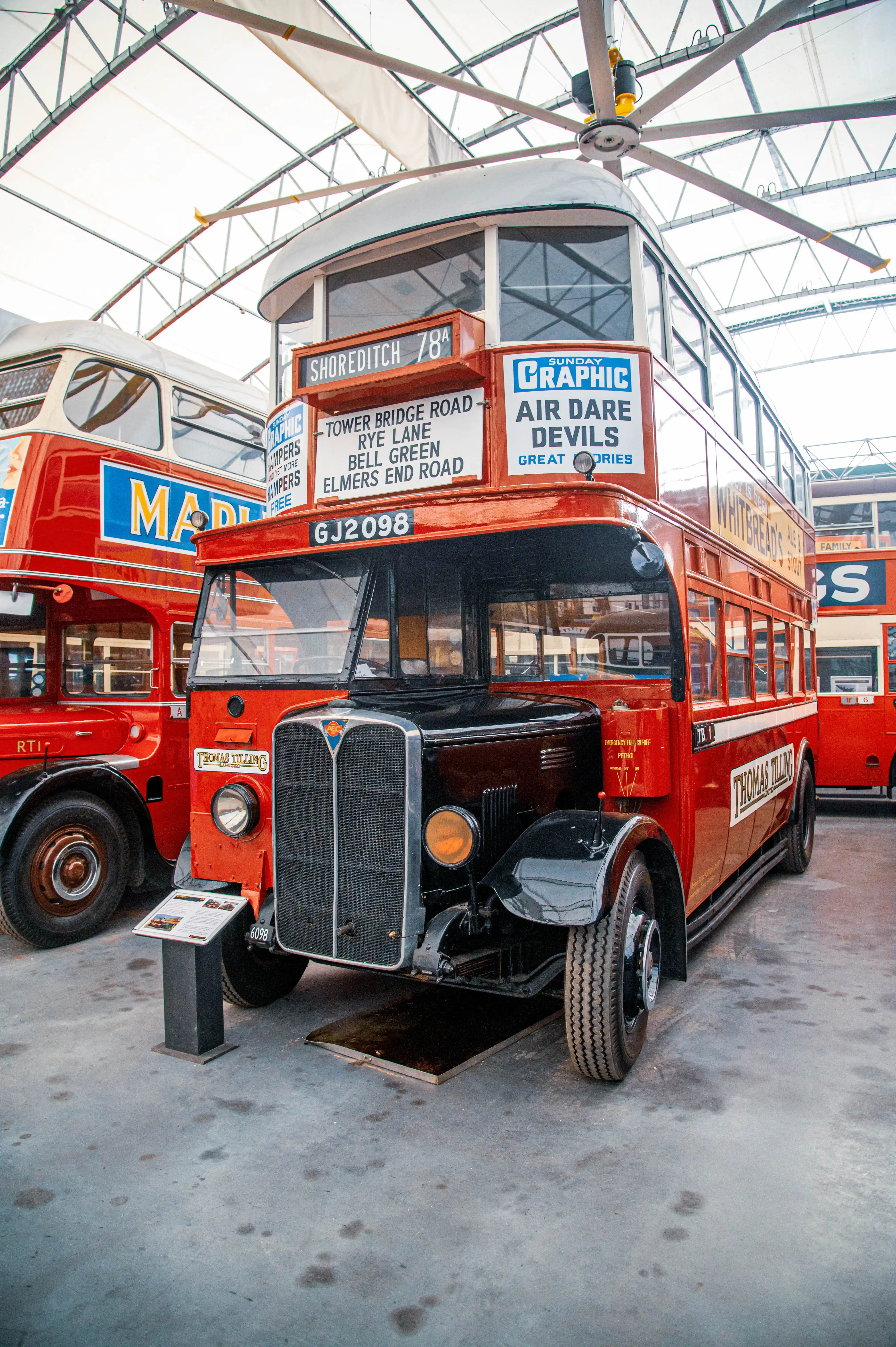 A close-up, angled view of a vintage red and black double-decker bus. The bus has signs on its front indicating routes and advertisements. It is parked indoors under a large industrial ceiling fan, with another similar bus visible to its left.