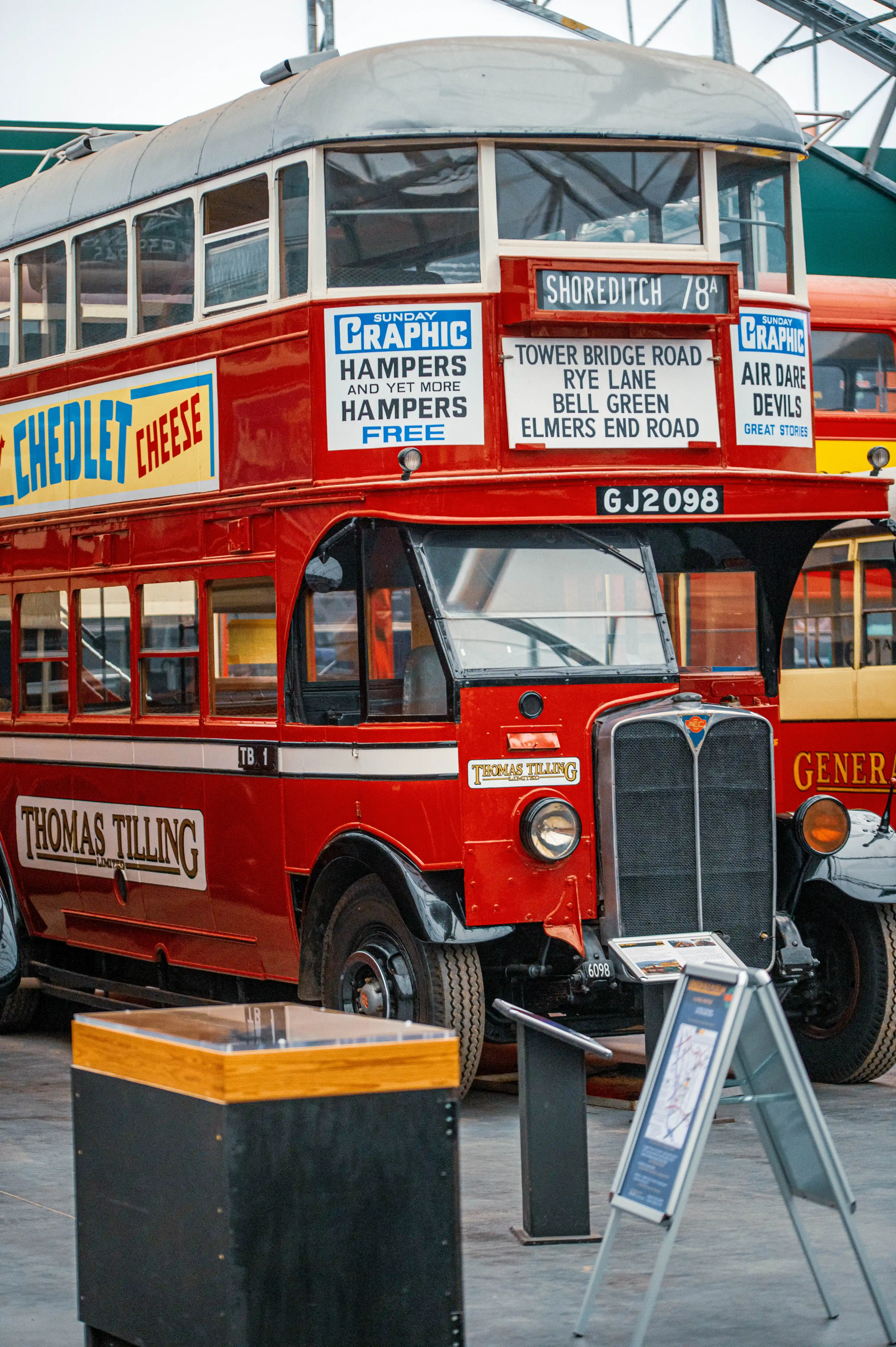 A bright red vintage double-decker bus with advertisements for Chedlet Cheese and Sunday Graphic Hampers, and route information displayed on the front.
