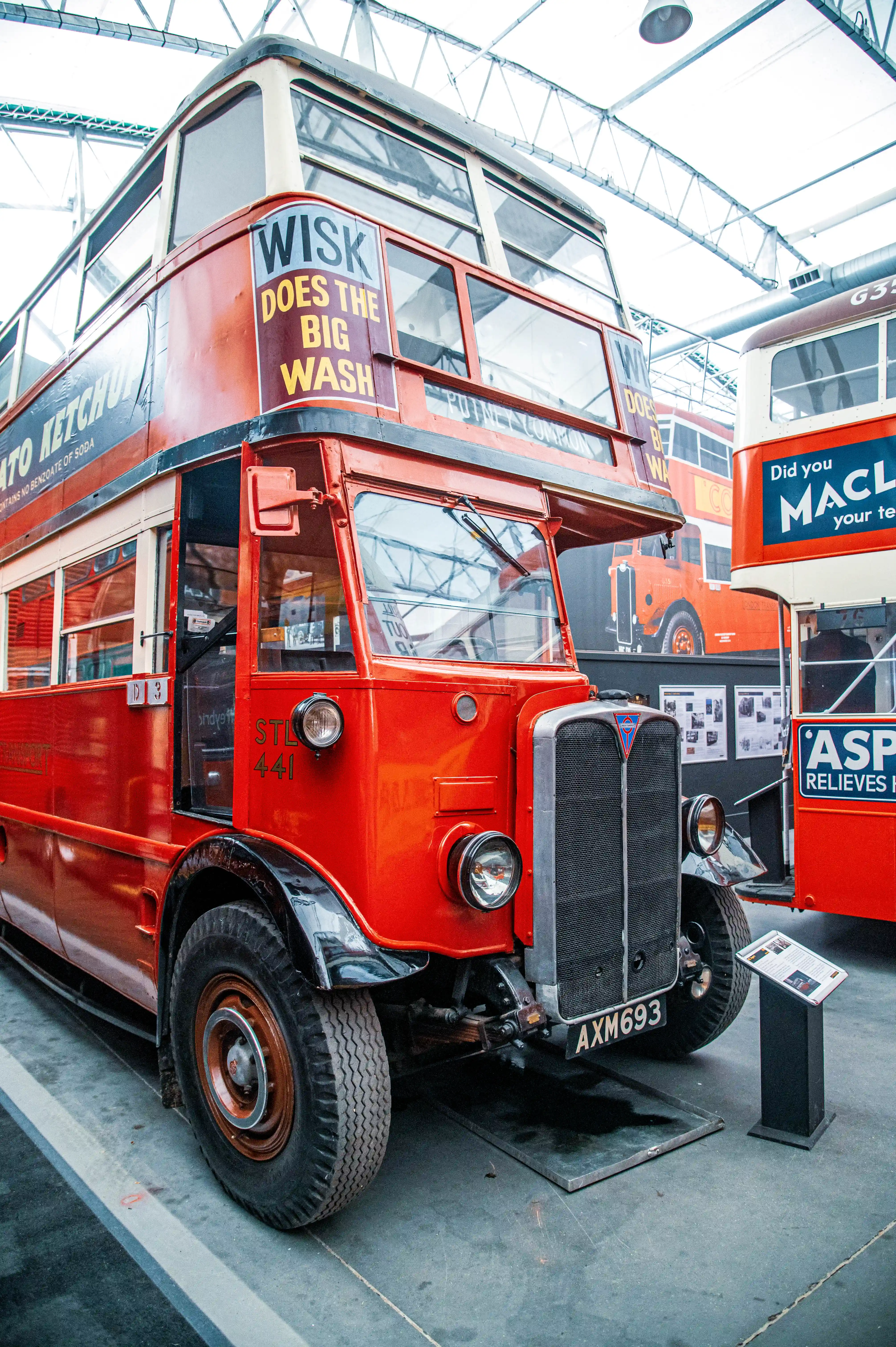 Close-up of a vibrant red vintage double-decker bus, viewed from the front corner. Advertising for WISK DOES THE BIG WASH is visible on the upper deck, and TOMATO KETCHUP on the lower deck. Another red bus with advertising for 'Did you MACLA... your te' is partly visible to the right.