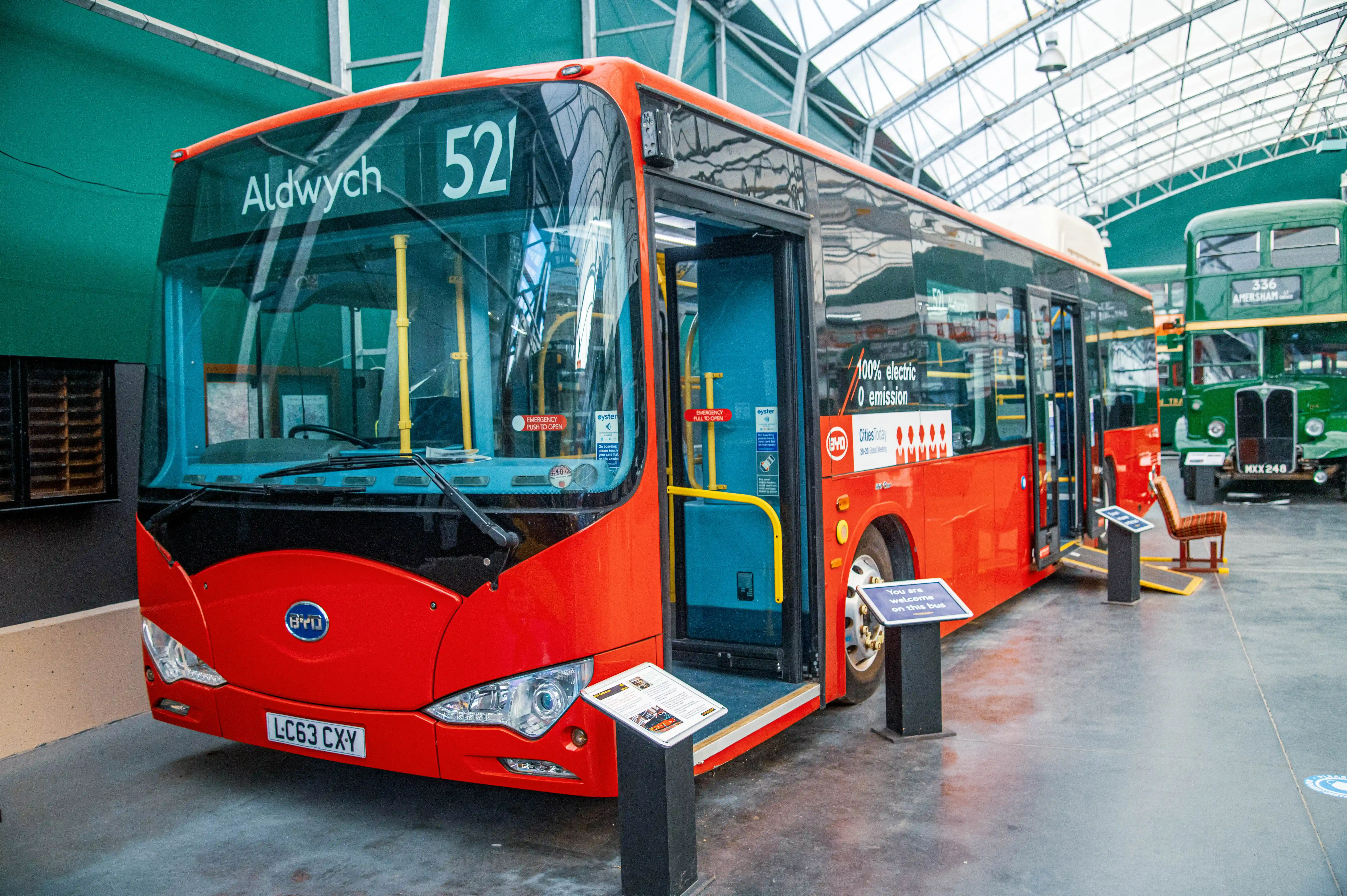 A modern red electric bus with route number 52 to Aldwych is shown with its front doors open, next to a vintage green double-decker bus in a museum setting.