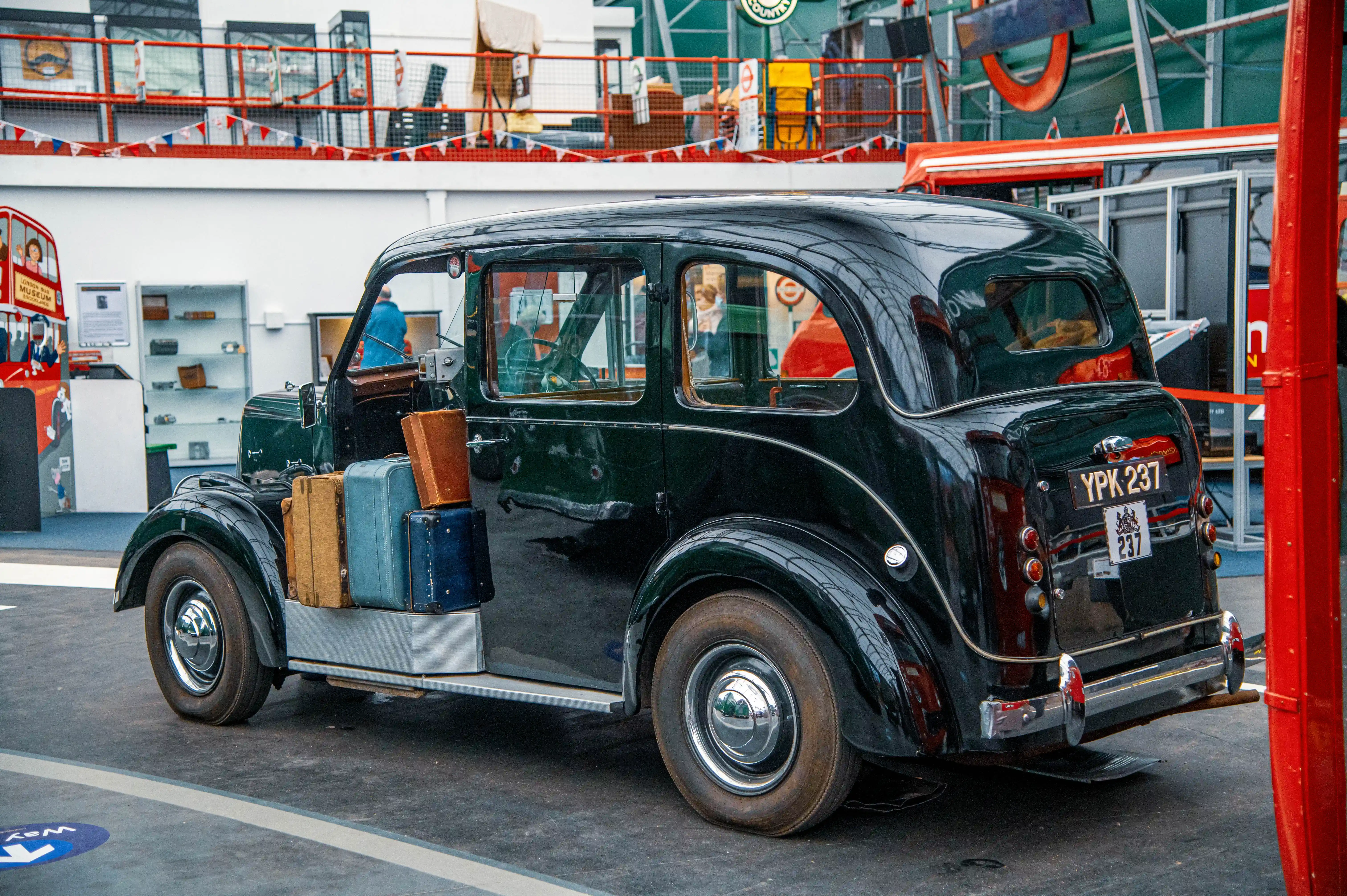 Side view of a vintage dark green London taxi with several old-fashioned suitcases stacked on its side, parked indoors.