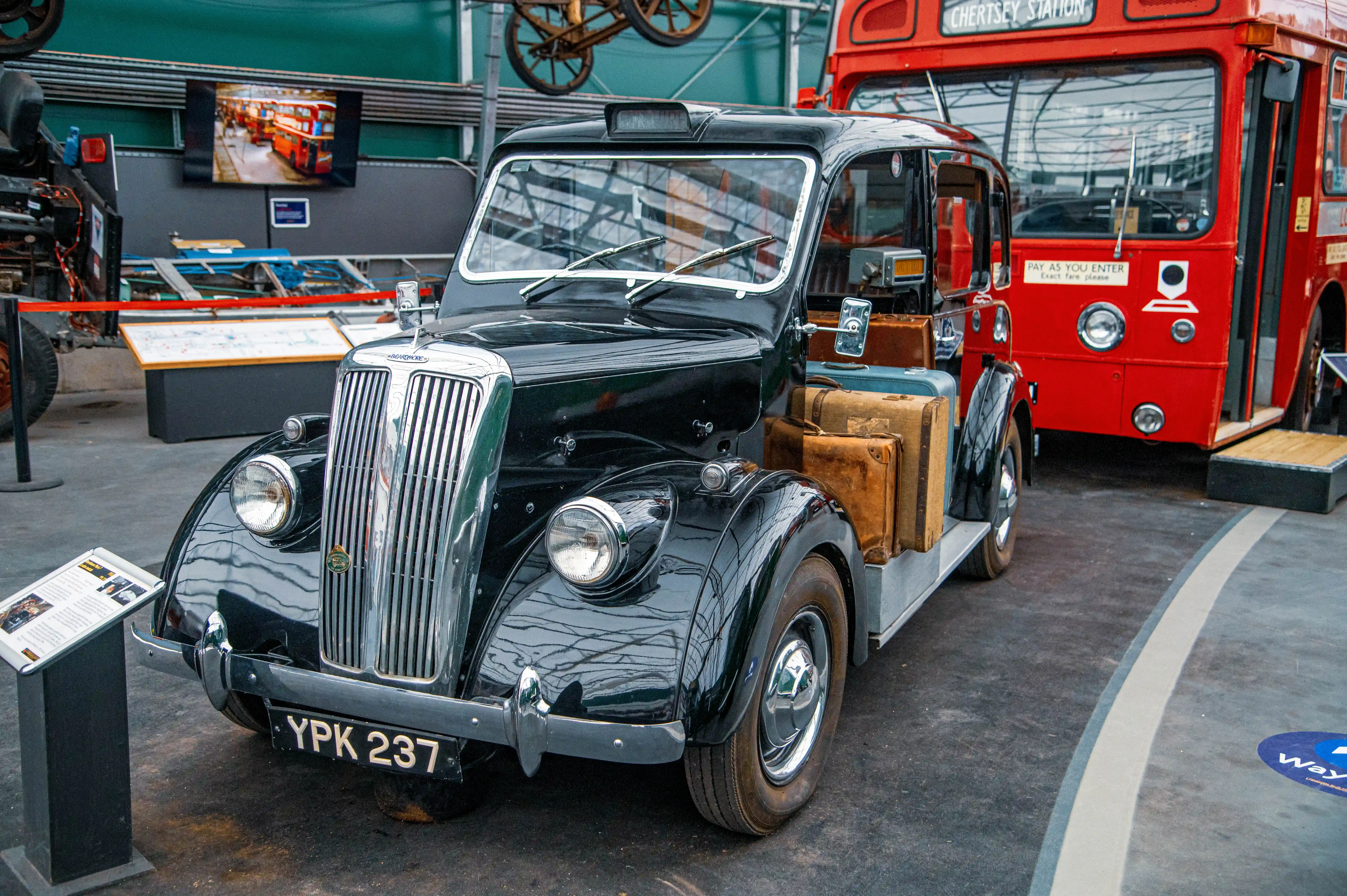 A vintage black London taxi, featuring a shiny chrome grille and period luggage stacked to its side, is parked next to a bright red Routemaster double-decker bus in an indoor exhibition.
