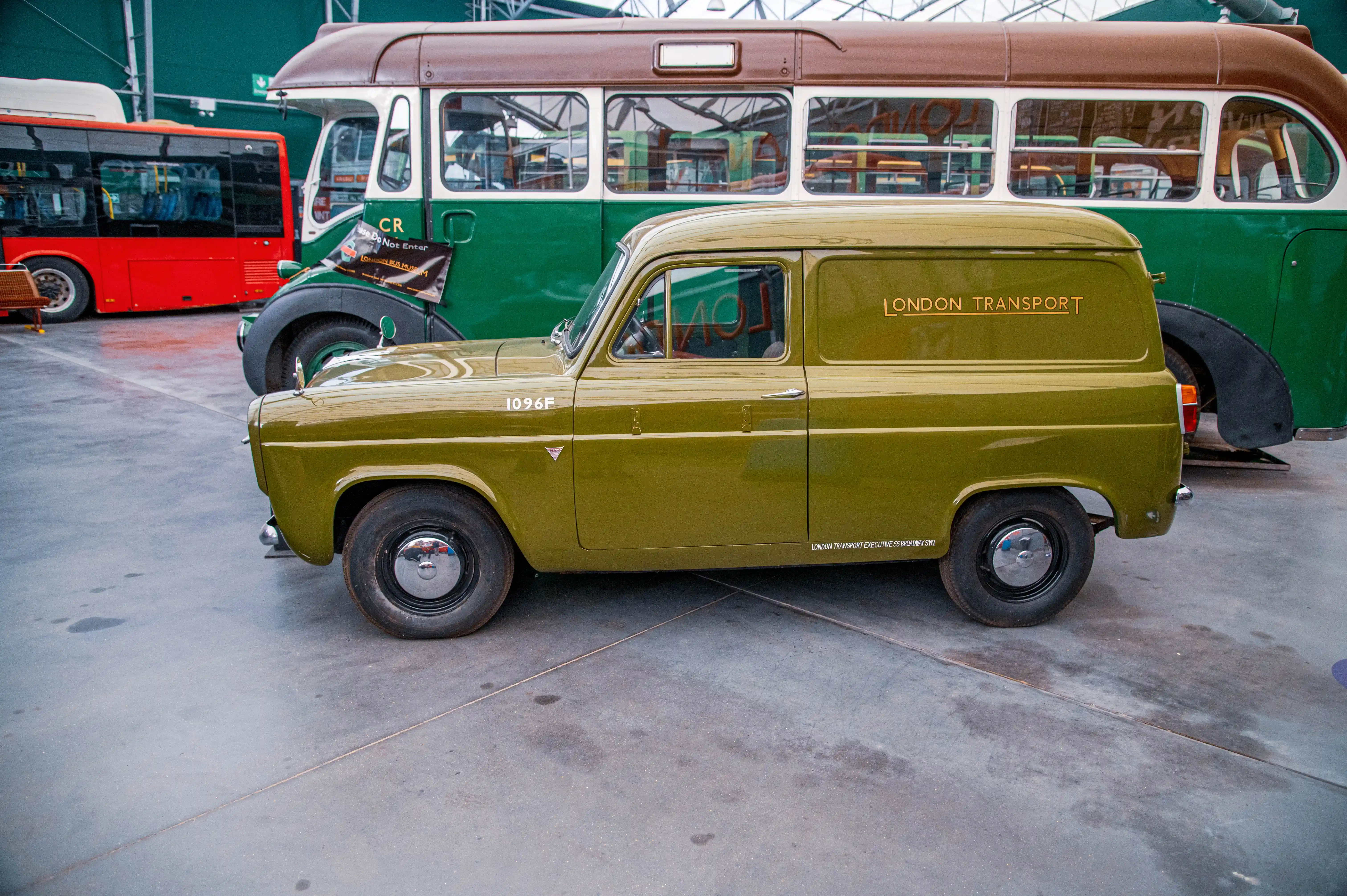 Side profile of an olive-green vintage van with 'LONDON TRANSPORT' written on its door, parked next to a green and brown vintage bus.