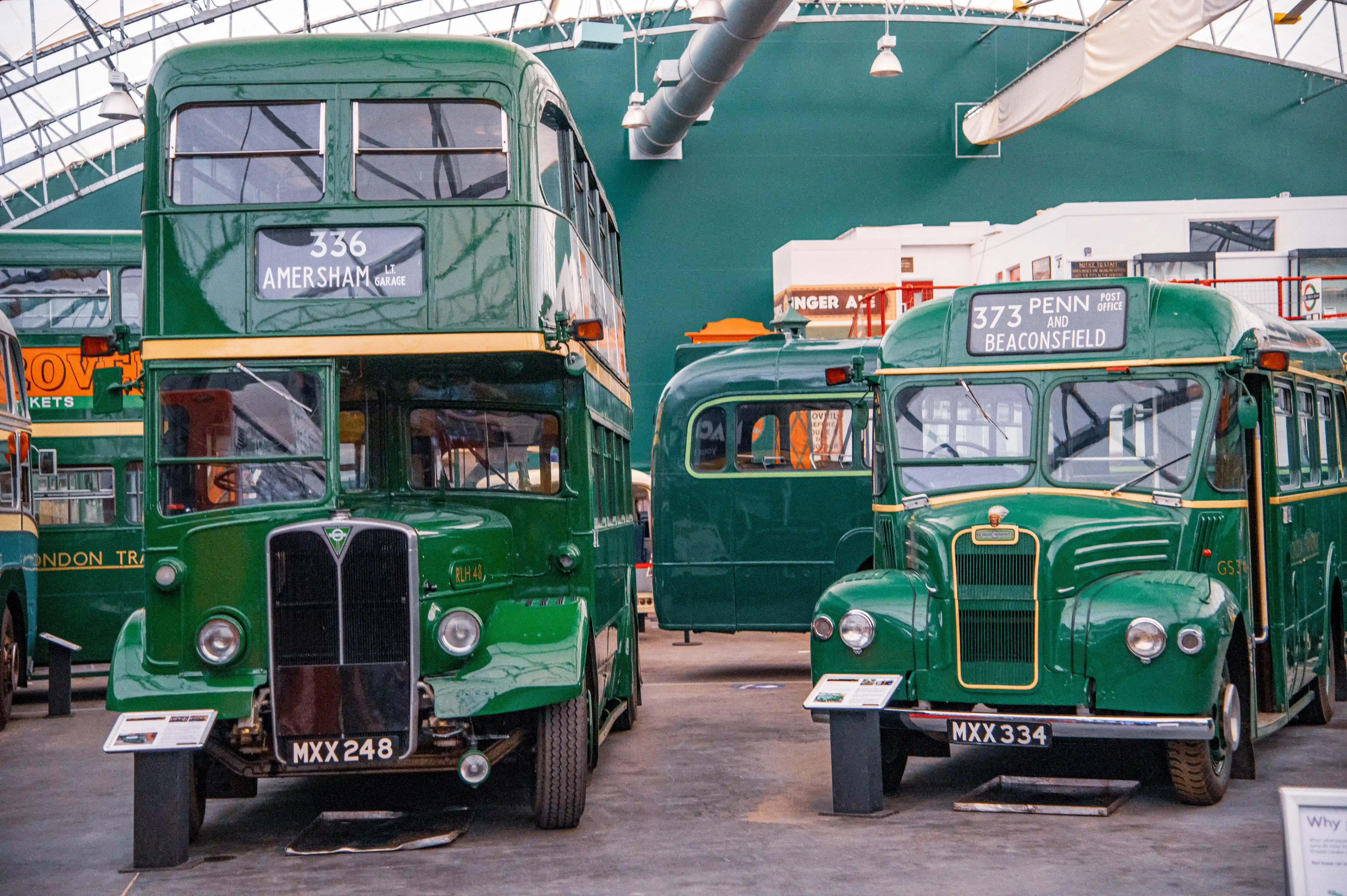 Two vintage green London buses, a double-decker on the left and a single-decker on the right, are parked side-by-side in a museum hall. The double-decker displays route number 336 to Amersham, while the single-decker shows route 373 to Penn and Beaconsfield.
