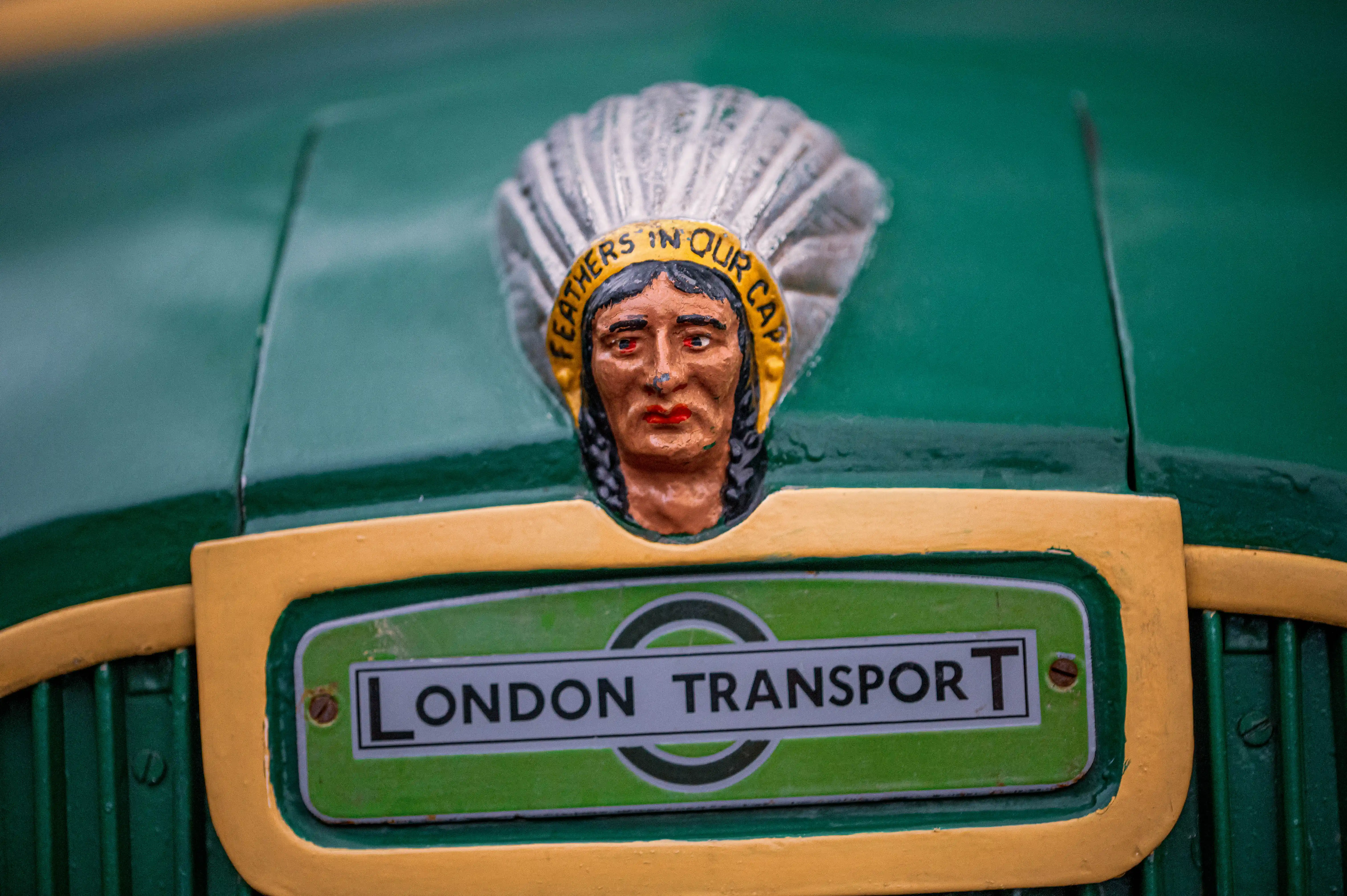 A close-up shot of a vintage London Transport emblem, prominently displaying a stylized head of a Native American wearing a feather headdress. The inscription 'FEATHERS IN OUR CAP' is visible on the headdress. Below this, the classic 'LONDON TRANSPORT' logo is featured within a green and yellow border.