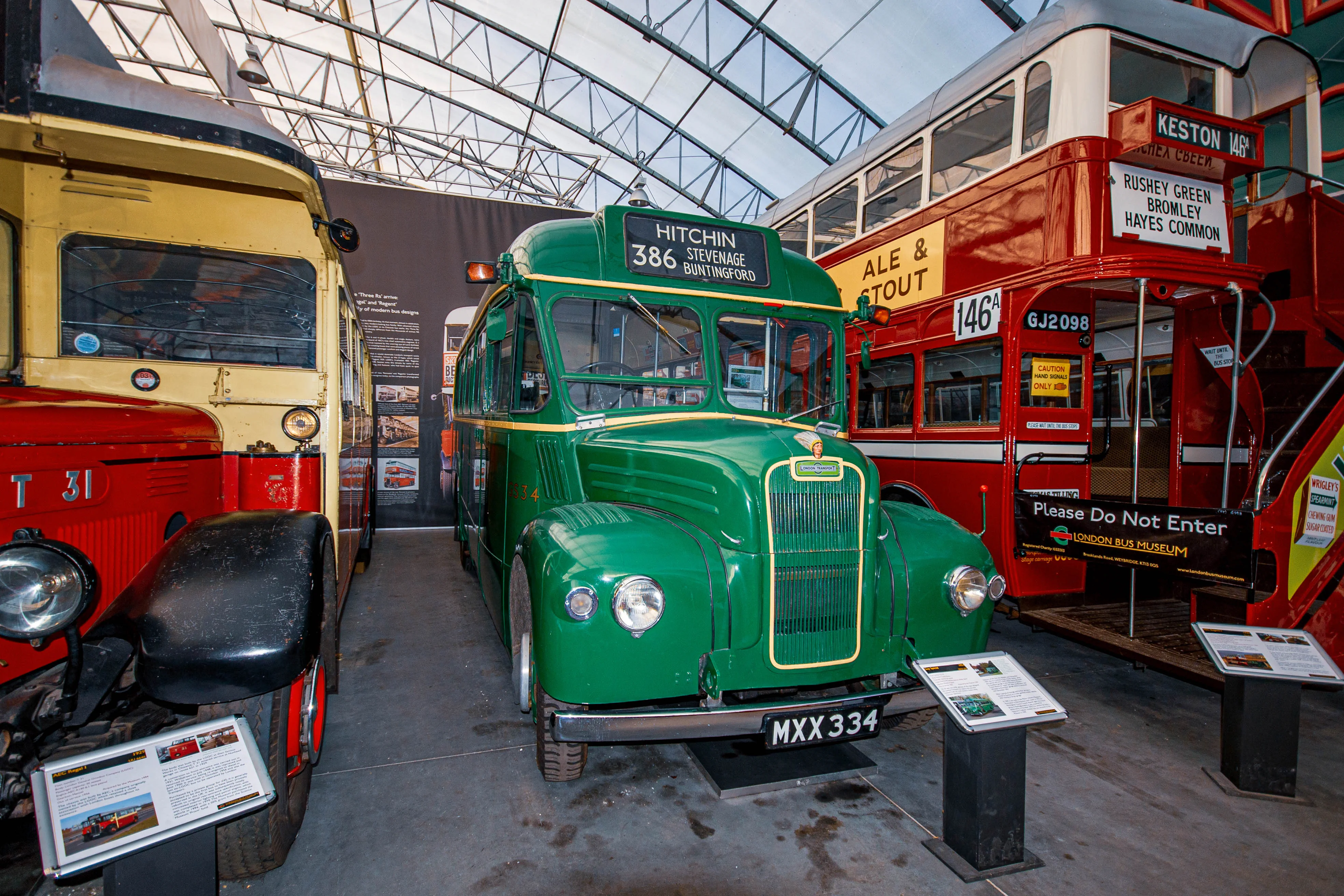 A museum display featuring a bright green single-decker bus parked next to a red double-decker bus, all under a modern translucent roof structure.