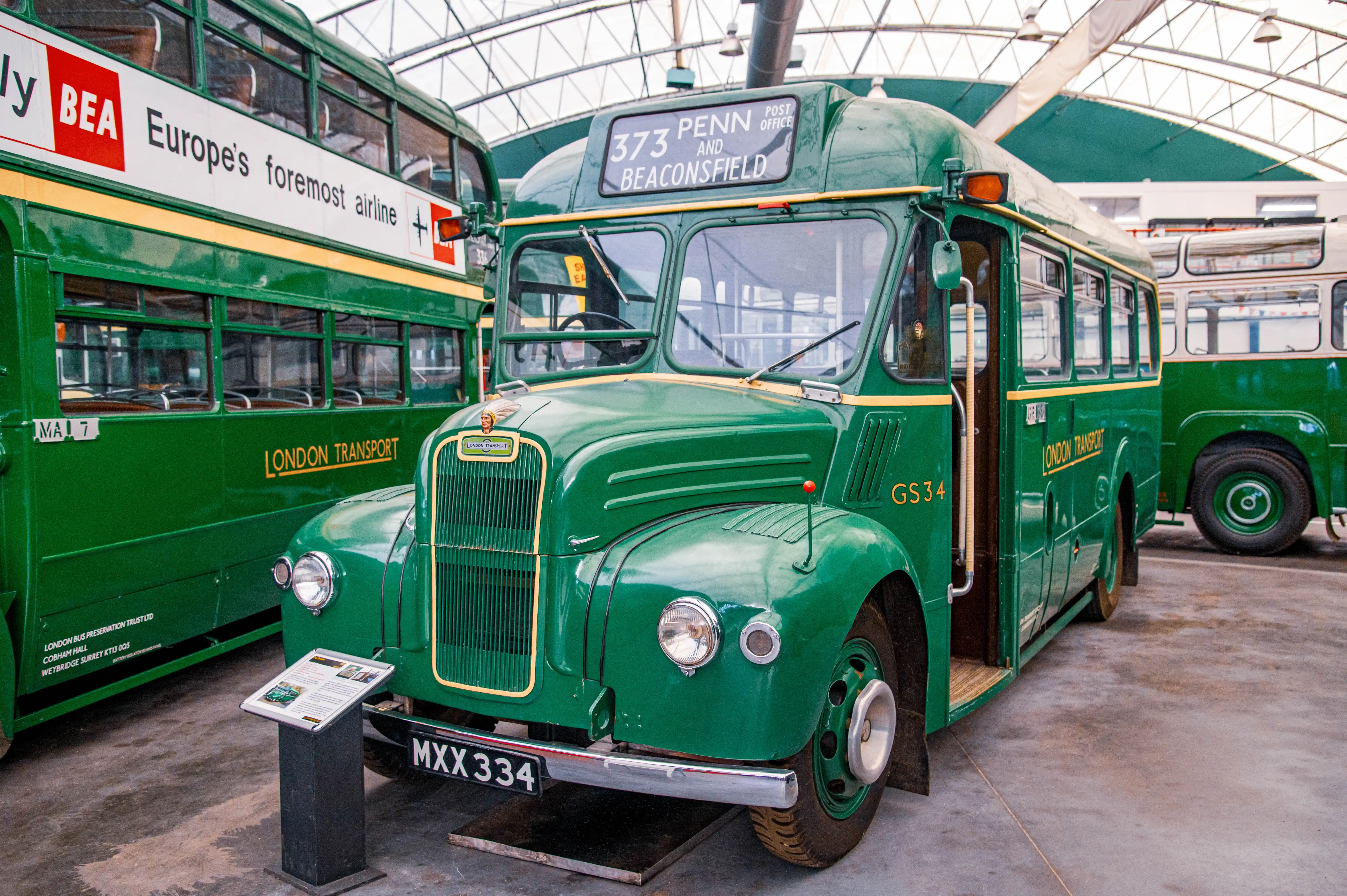 A close-up of a vintage green London bus, marked GS34 and MXX 334, with another green double-decker bus featuring a Europe's foremost airline advertisement partially visible to the left.