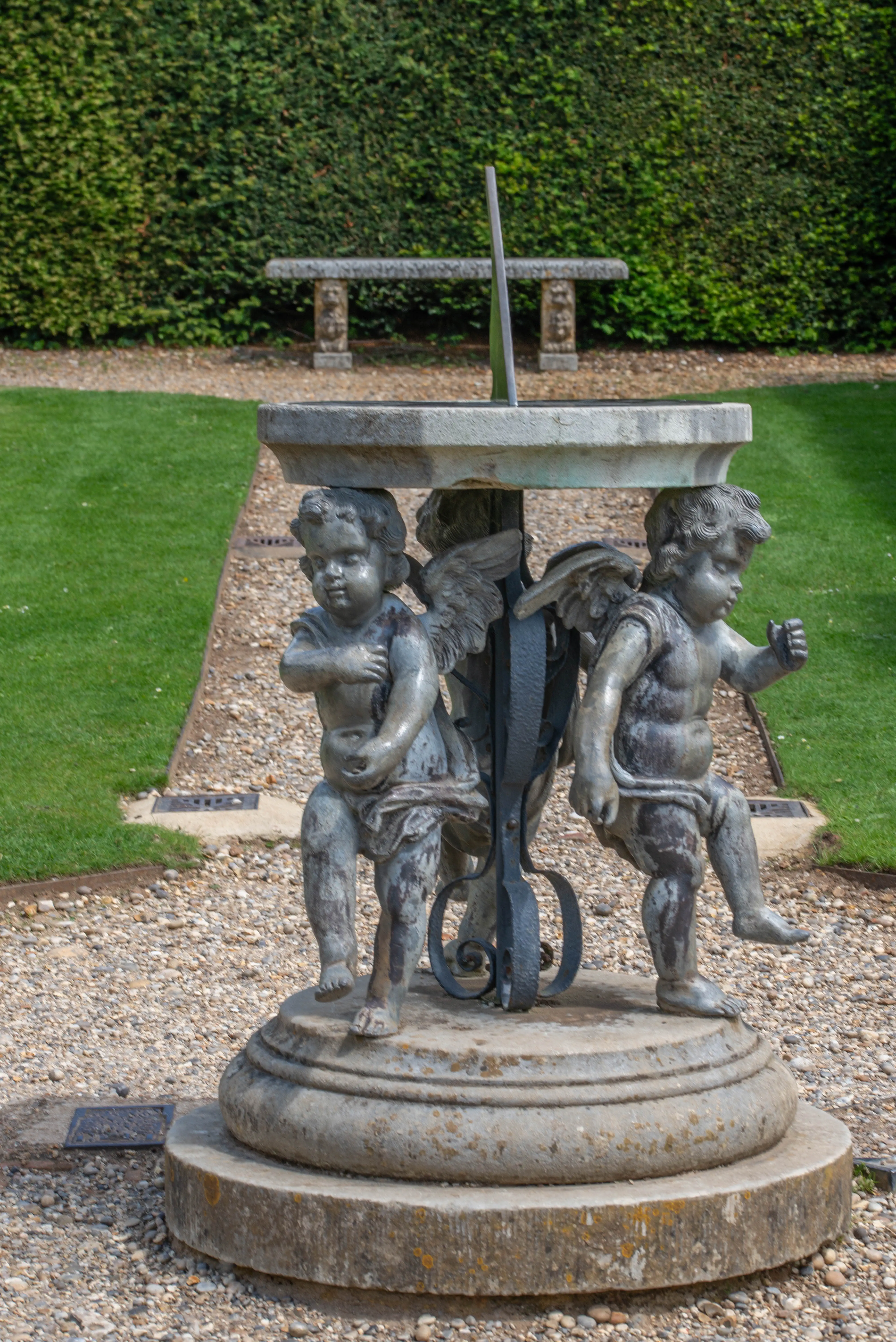 Stone sundial with cherub statues as support, set in a garden with a hedge and lawn.