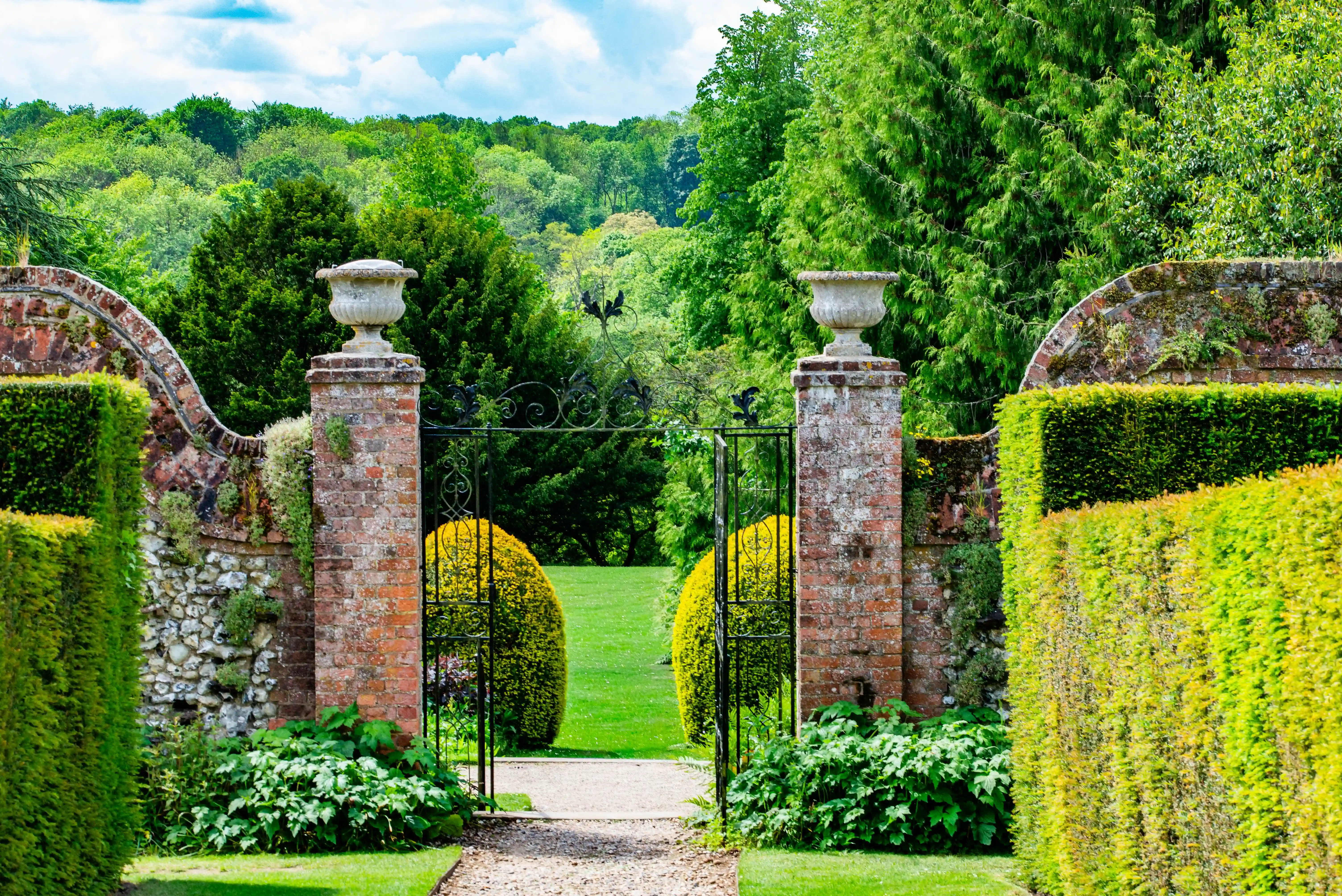 A garden path leads to ornate black iron gates set between brick pillars and stone walls, opening to a view of bright green shrubs and a lush woodland in the distance.