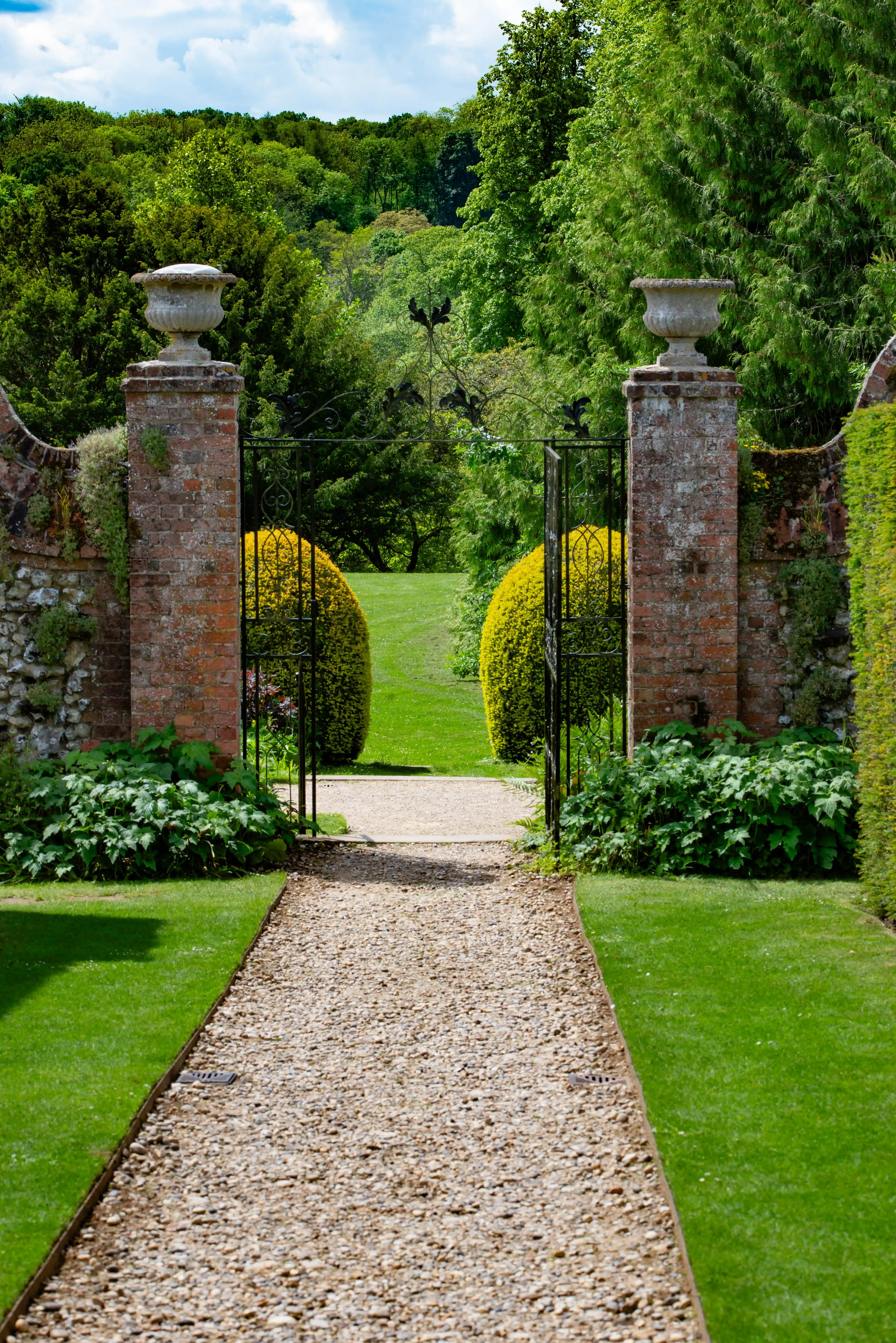 Gravel path leading through decorative brick pillars and wrought-iron gates into a bright green English country garden with rolling hills in the background.