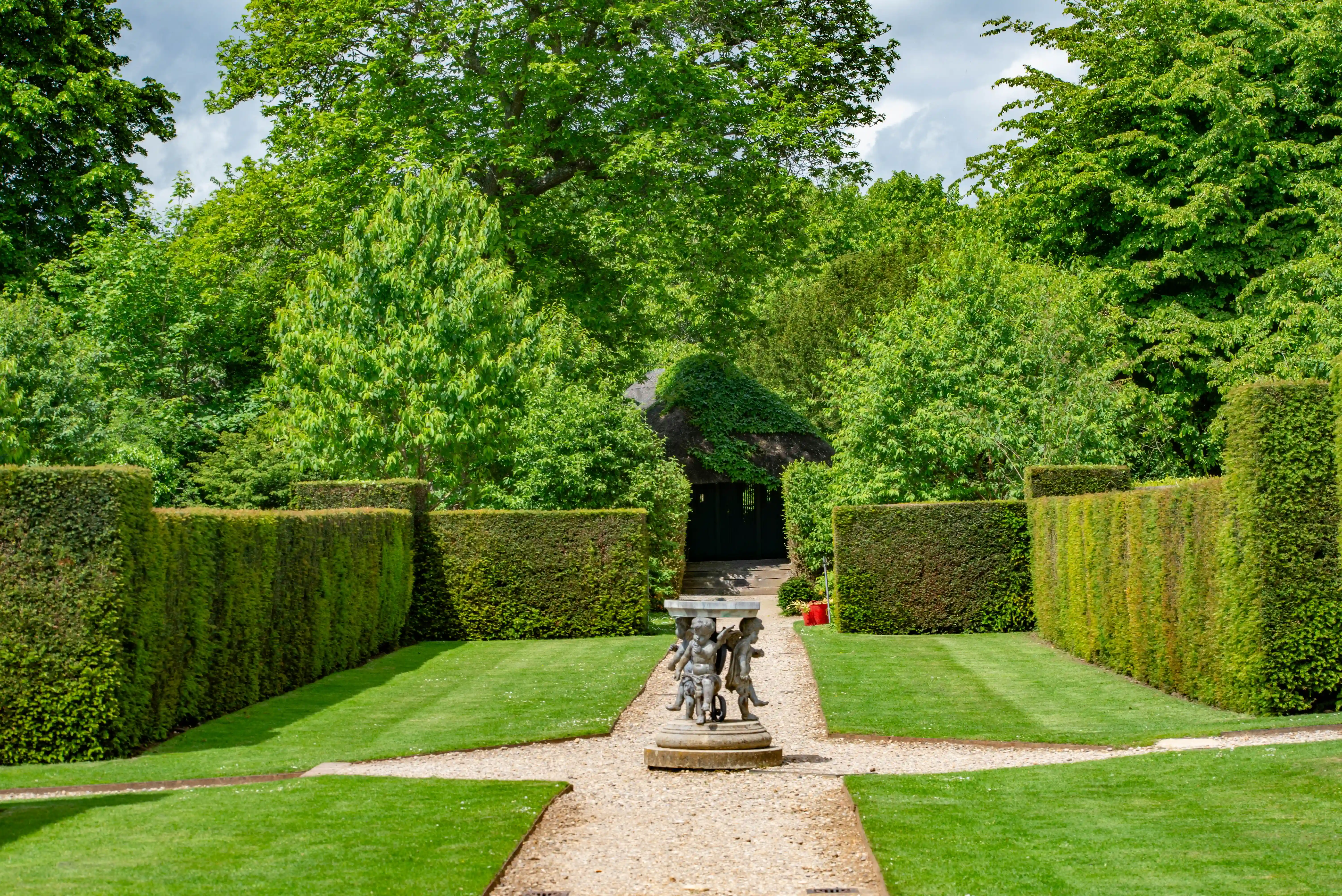 A symmetrical garden path directs the eye towards a stone fountain depicting cherubs, with tall, clipped hedges on either side. Beyond, a thatched building peeks through dense green foliage.