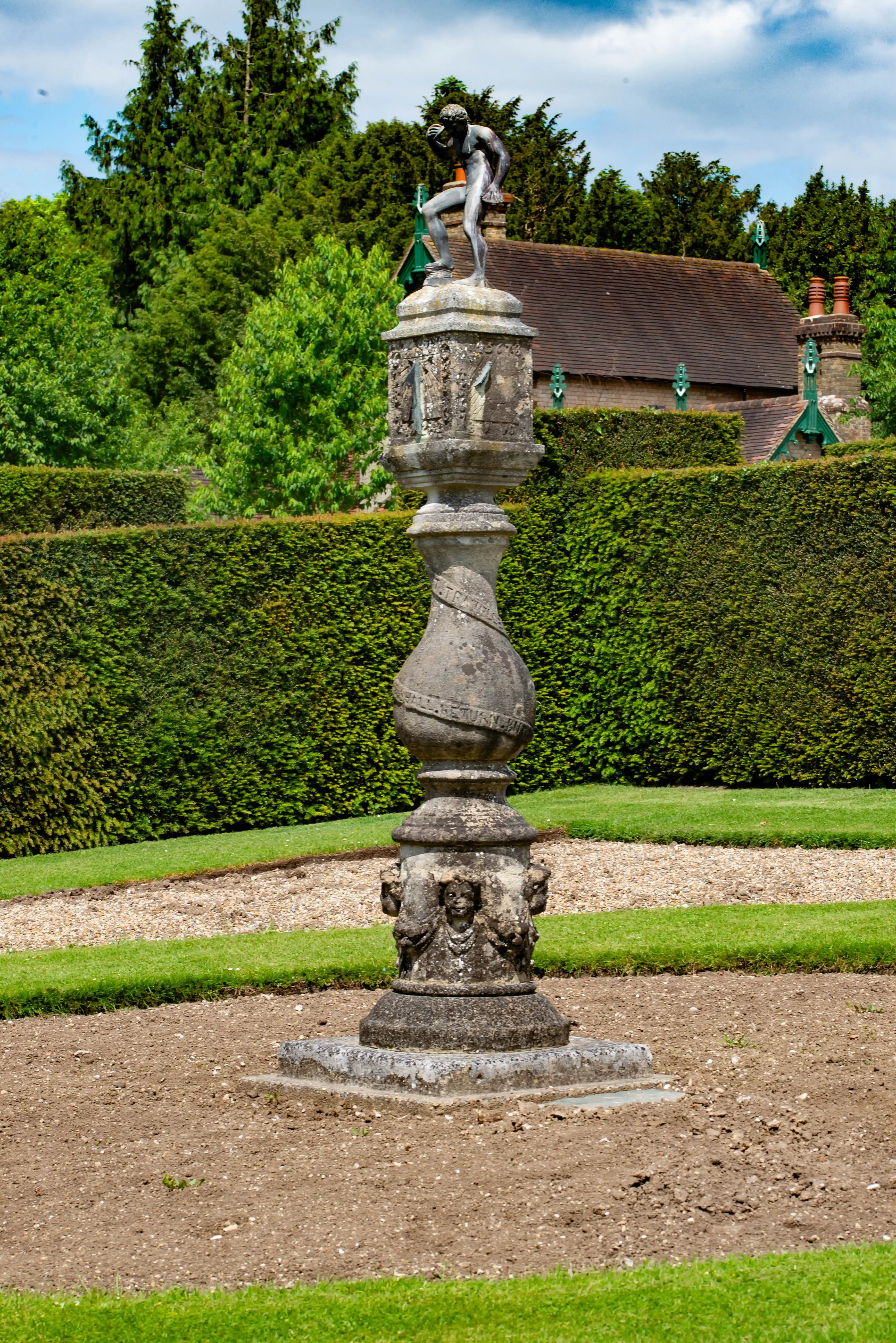 A stone sundial with a bronze statue stands in a garden bed, with a tall green hedge behind it and a building and trees in the distance.