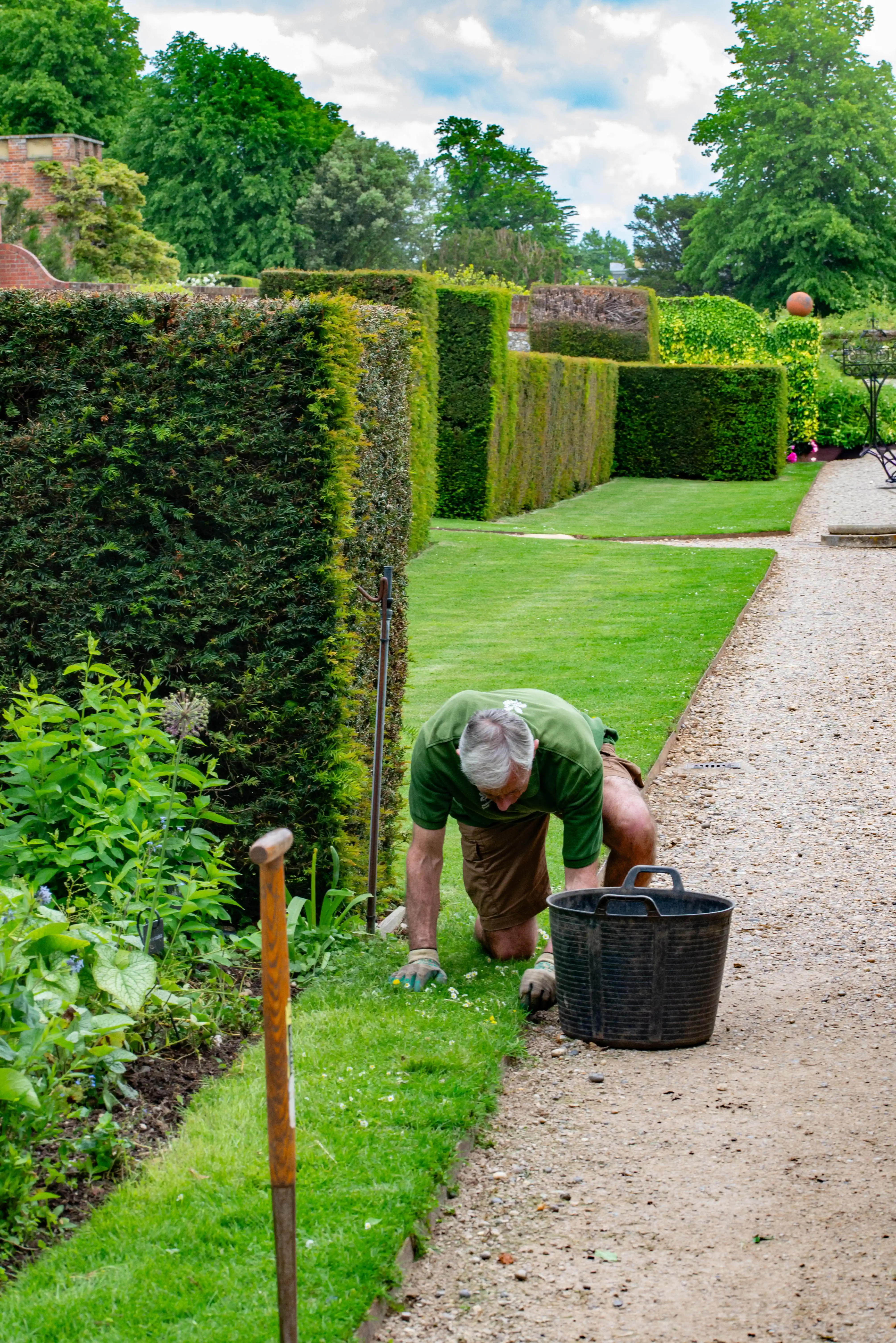 A gardener kneels by a gravel path in a formal garden, working on the lawn edge.