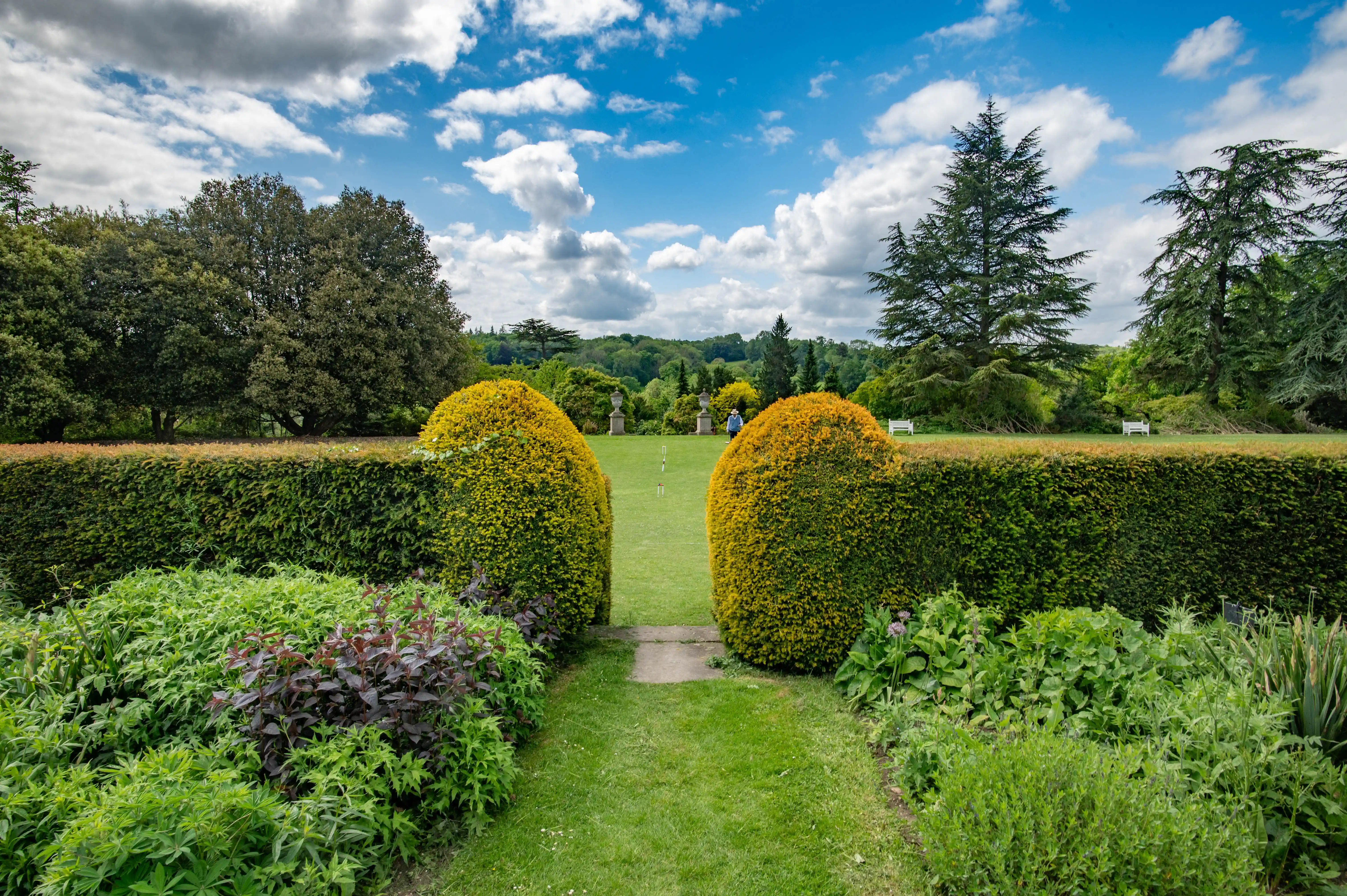 A view through a garden opening, showing a well-manicured lawn, tall hedges, and a person playing croquet in the distance, with a backdrop of trees and a cloudy blue sky.