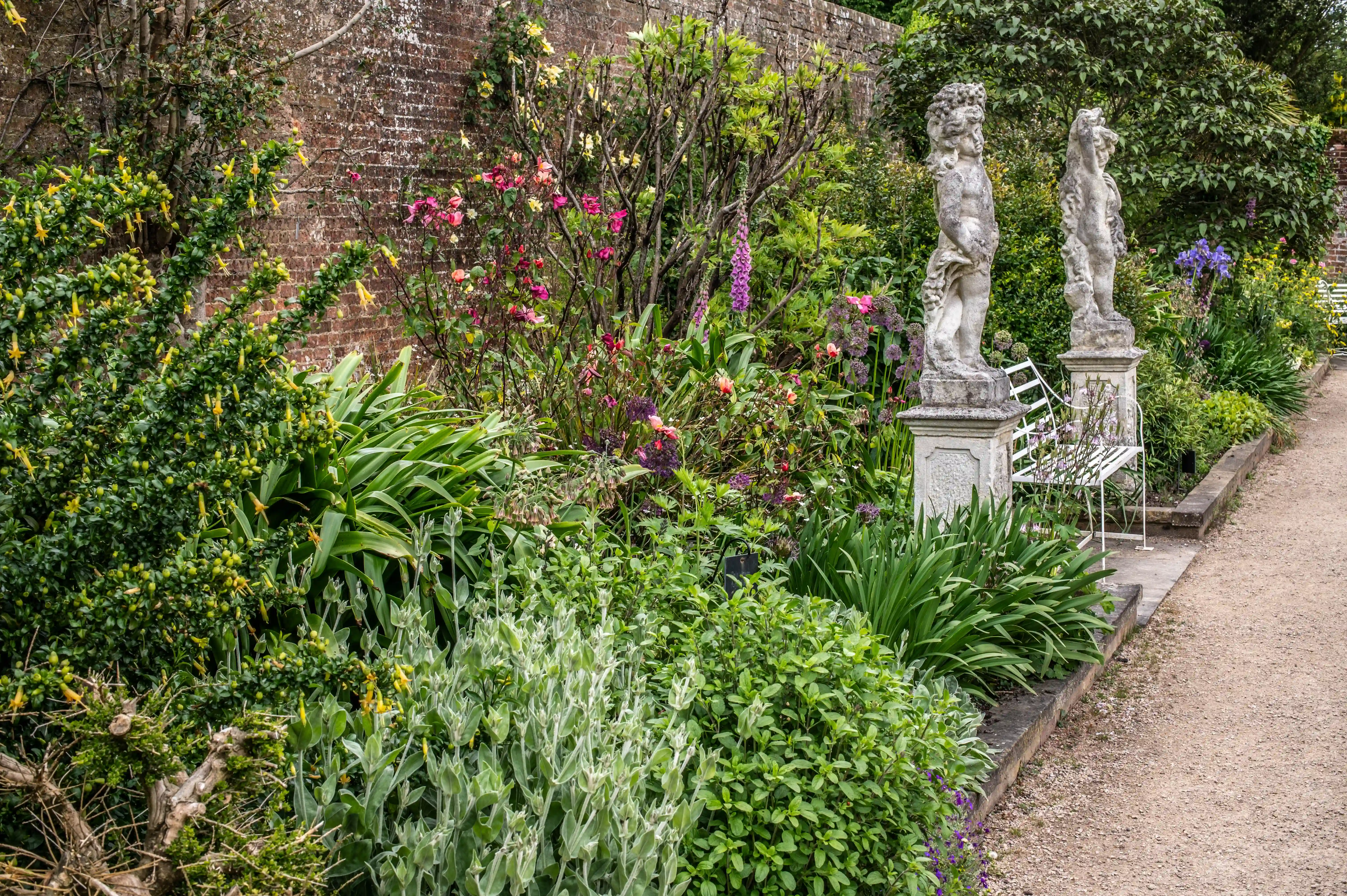 An image of a garden path with various flowers, plants, two stone statues, and a white bench.