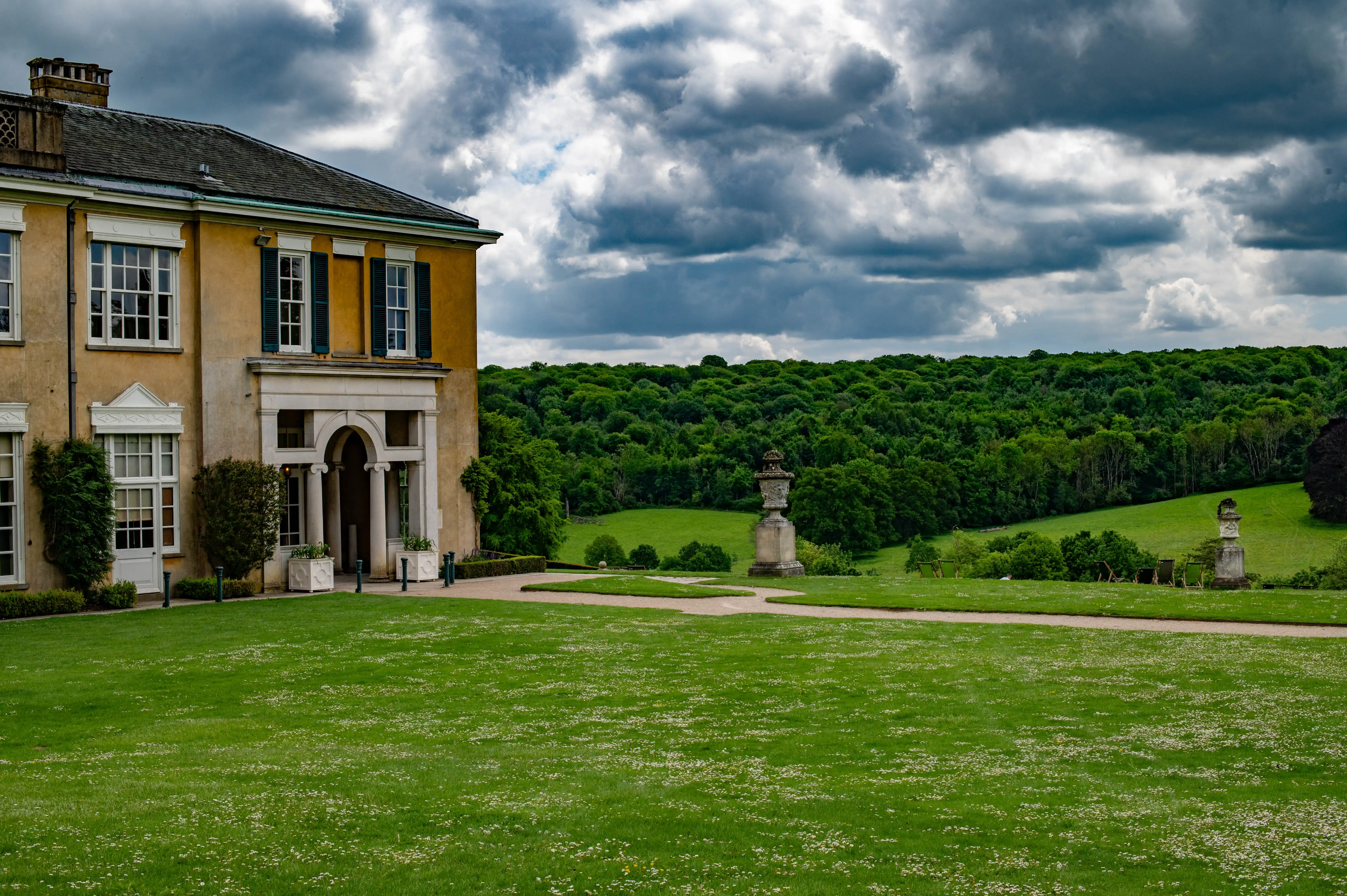 A wide shot of a grand yellow country house with a portico, set within extensive parkland. A large, green lawn stretches towards a backdrop of dense, green woodland. The sky is filled with dramatic, dark grey clouds, with patches of lighter sky visible.