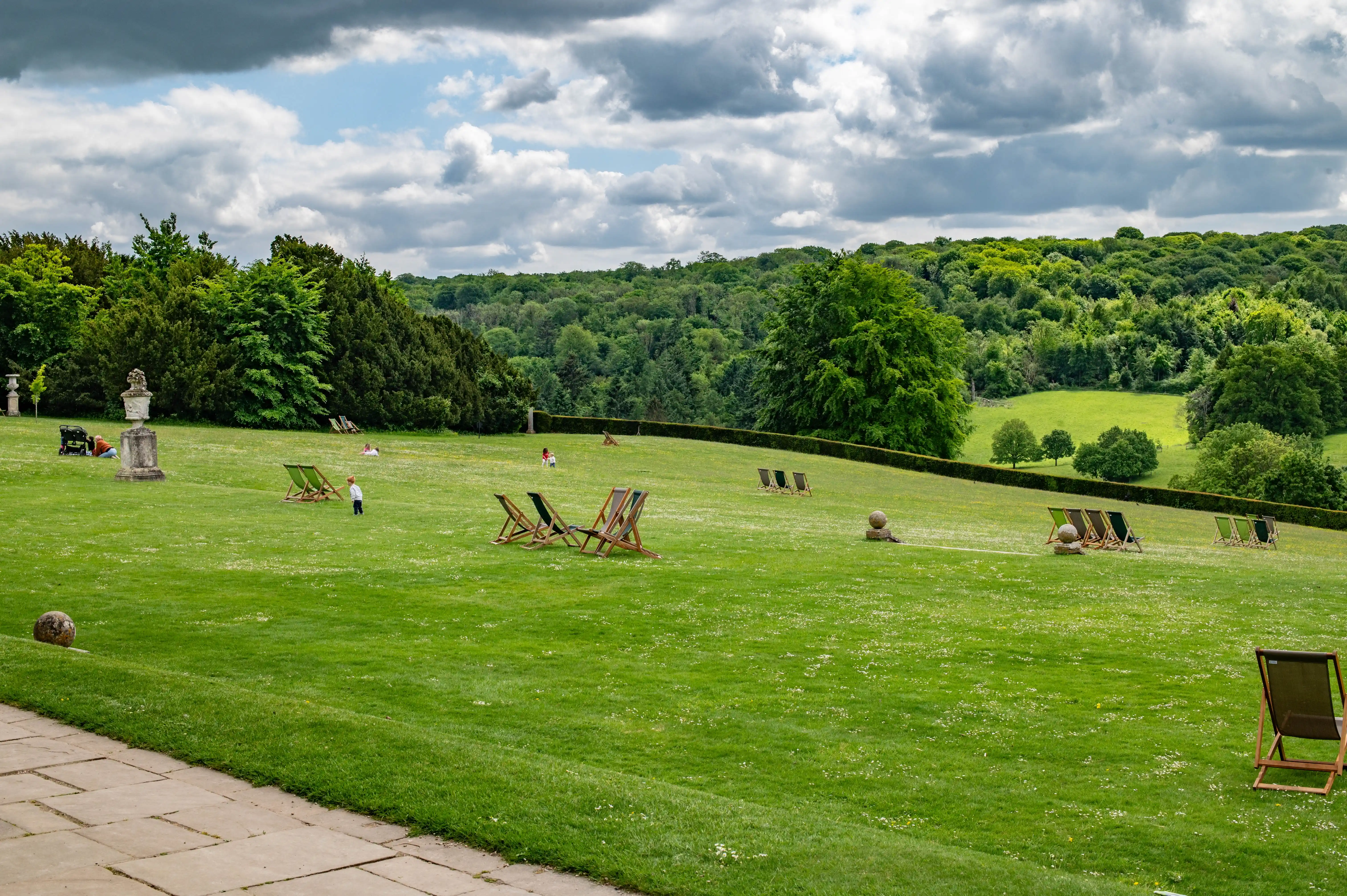 Wide shot of a large, green lawn in an English garden with white wildflowers, several empty deckchairs, a few distant figures, and a backdrop of lush, rolling hills and trees under a cloudy sky.