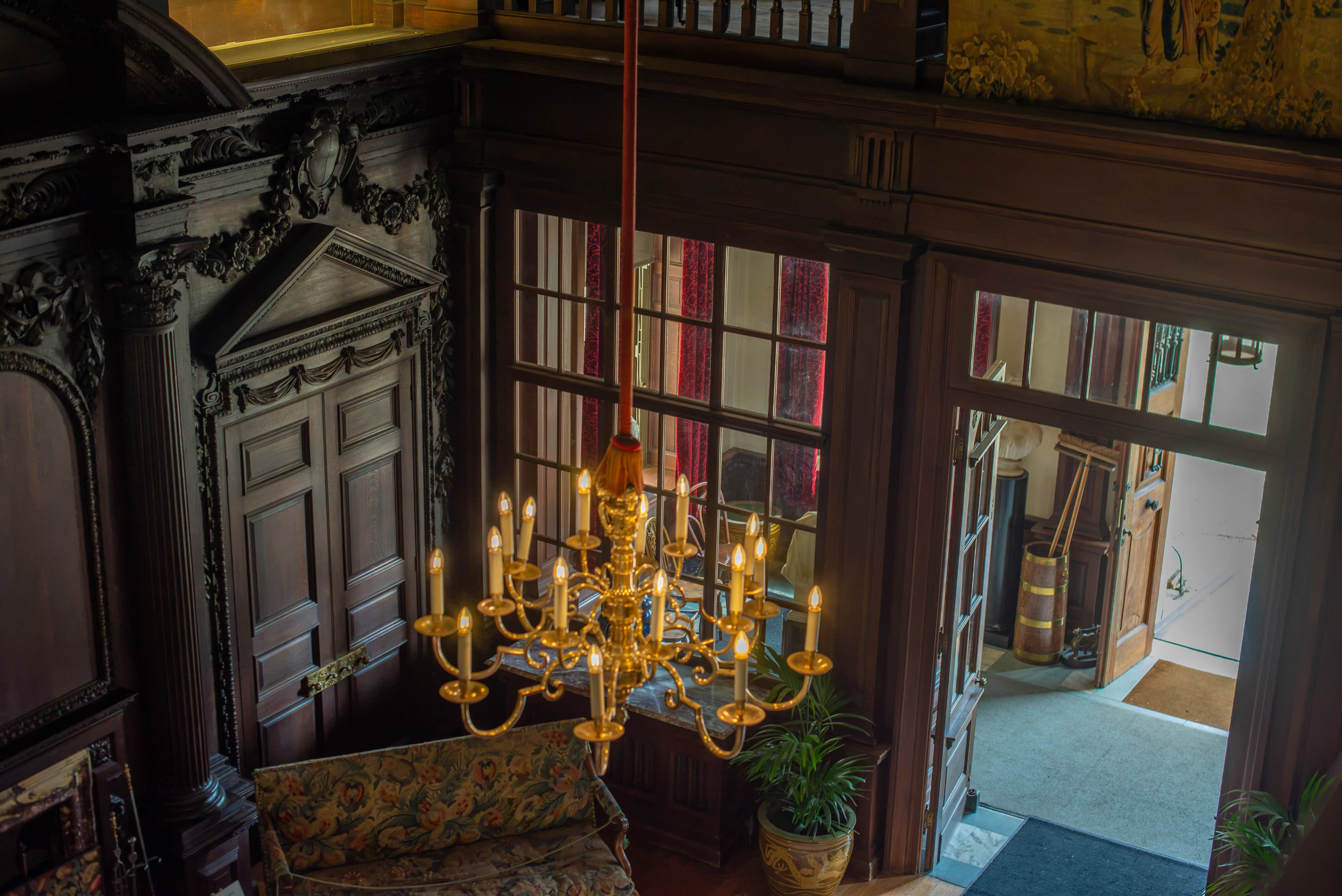 A top-down view of a grand hall with dark wood panelling, a large brass chandelier, a sofa with a floral pattern, and a view through a window and doorway..