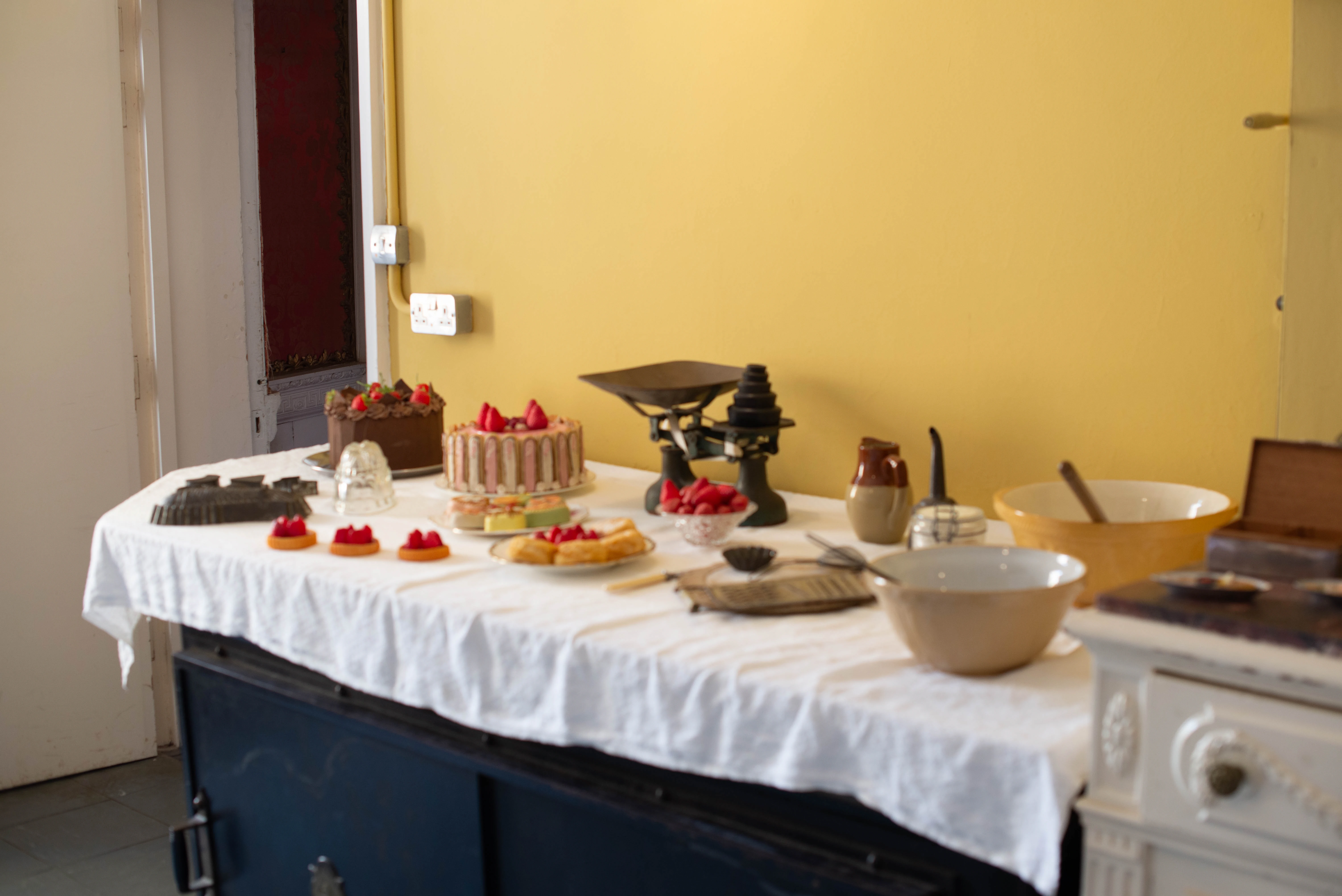 A close-up shot of a table laden with various baked goods, including a chocolate cake decorated with strawberries, a pink and white striped cake topped with strawberries, and several small tarts. Baking equipment such as scales and bowls are also visible.