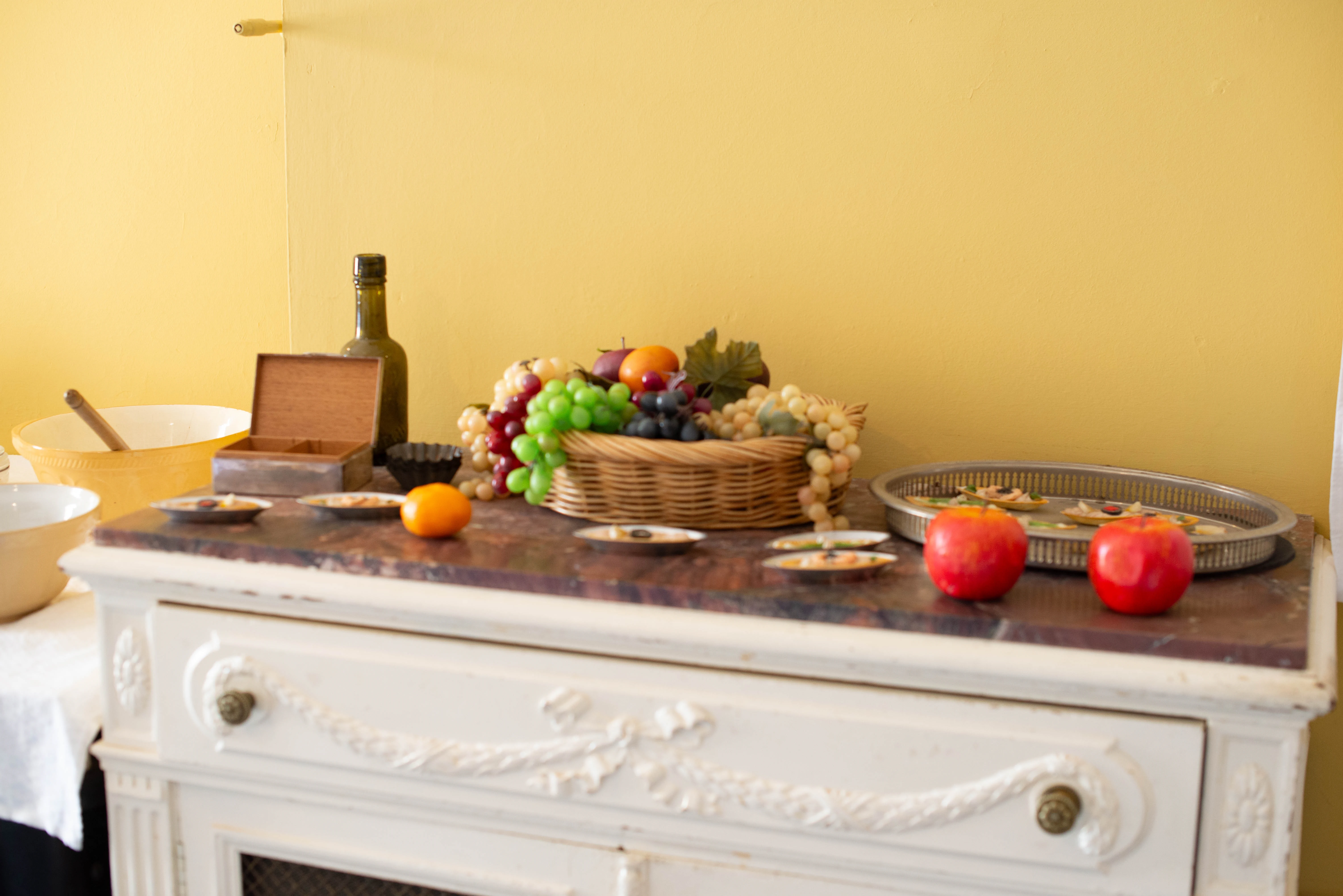 A still life photograph depicting a woven basket filled with red and green grapes, plums, and oranges, placed on a marble-topped dresser. Two red apples and a tray of small appetizers are also visible, with a yellow wall in the background.