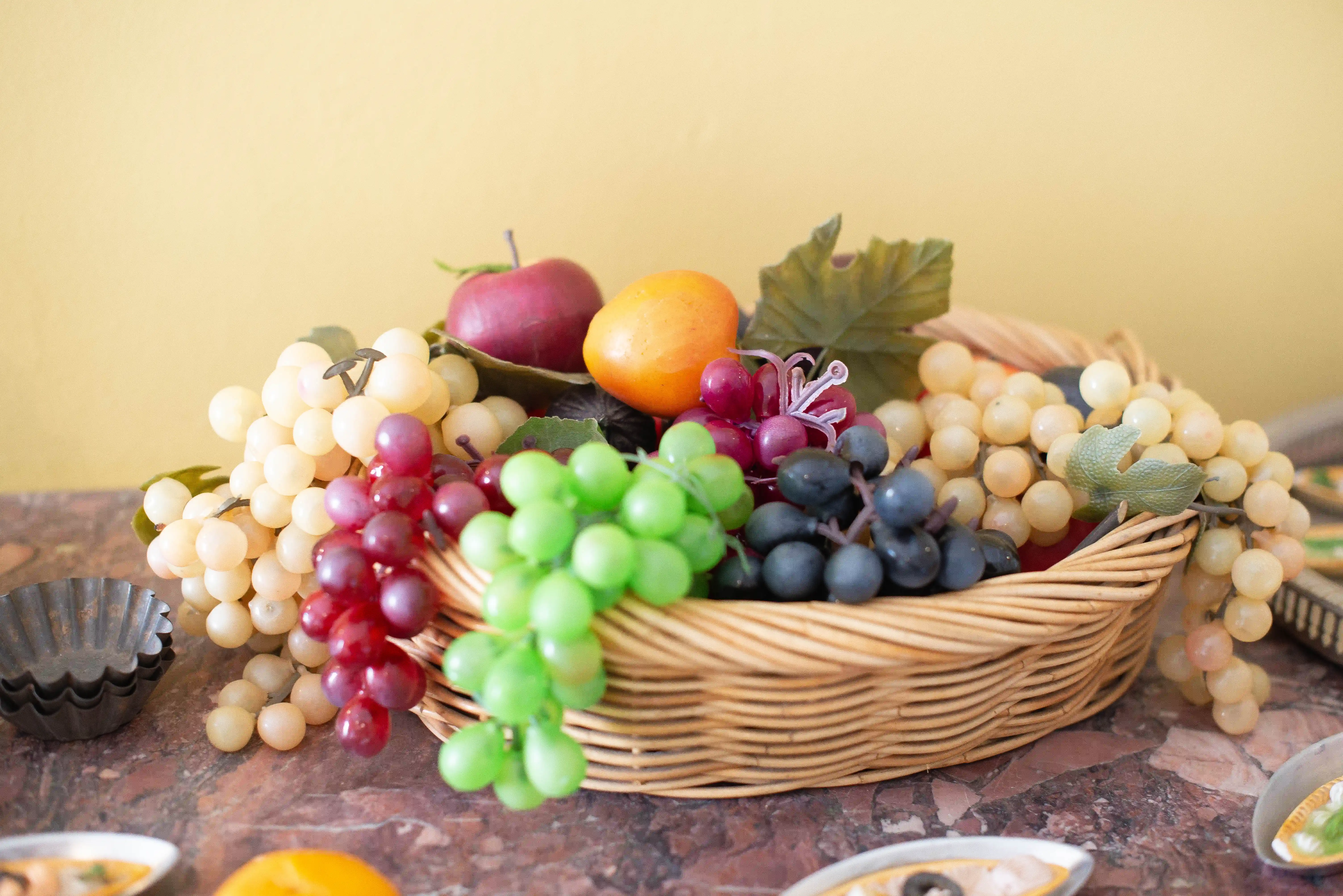 A woven wicker basket filled with bunches of artificial grapes in shades of cream, red, green, and dark purple. A red apple and an orange are nestled amongst the grapes, with artificial leaves providing a touch of greenery. The basket sits on a mottled marble surface, with the background a soft yellow.