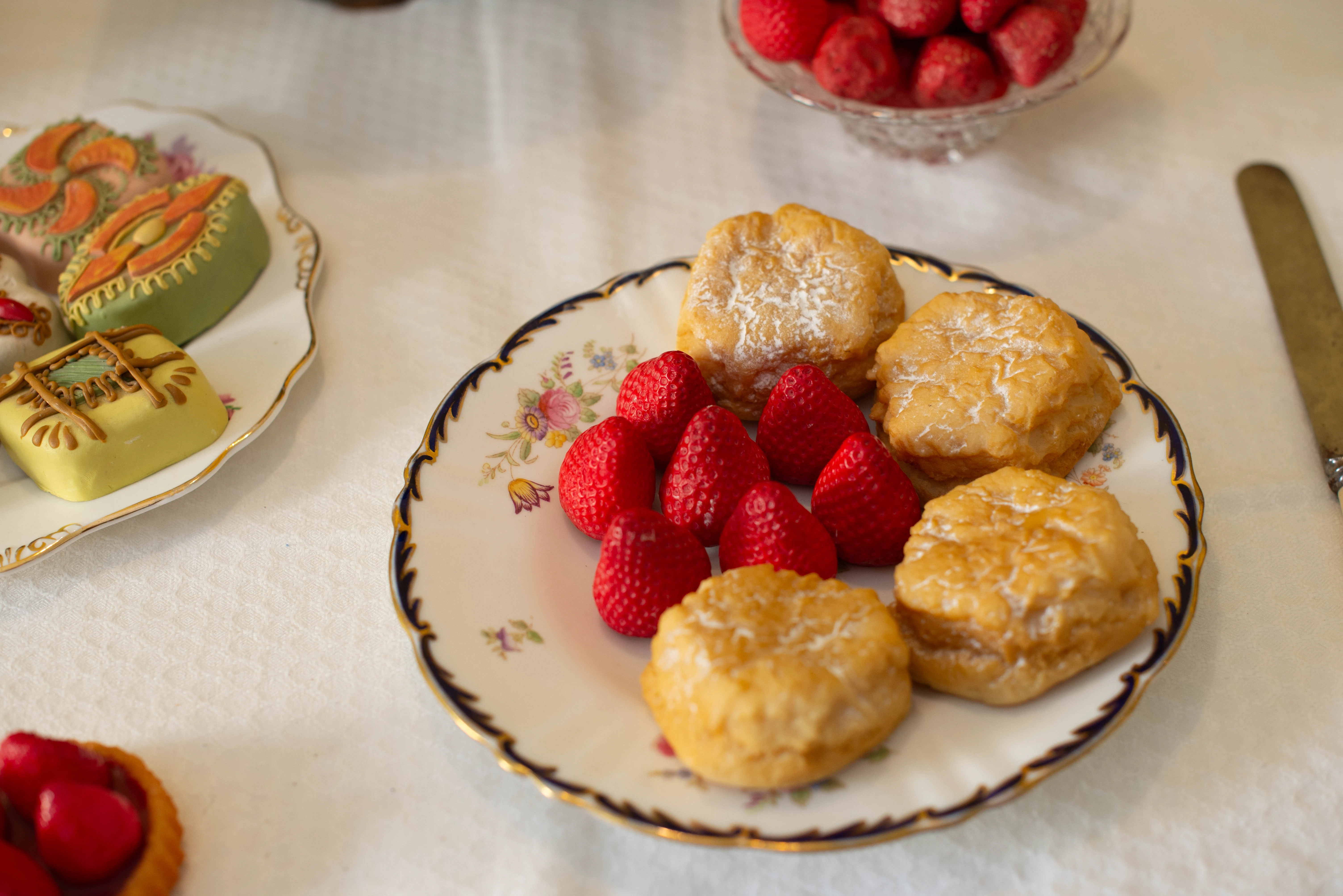 A close-up shot of a decorative porcelain plate filled with bright red strawberries and golden-brown scones dusted with powdered sugar, alongside a plate of artfully crafted petit fours.