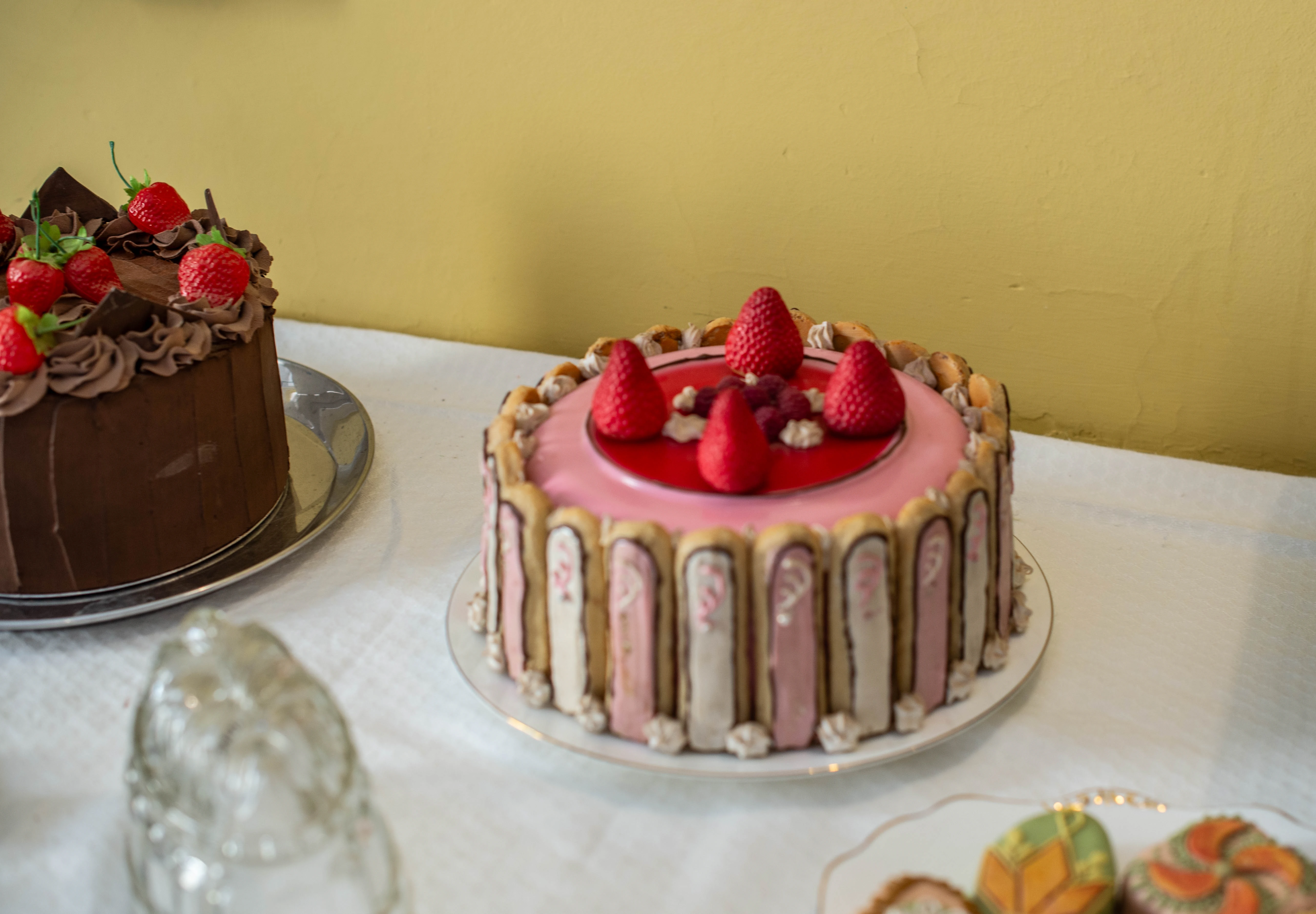A chocolate cake with chocolate frosting and strawberries sits to the left of a pink glazed cake decorated with biscuit fingers, strawberries, and raspberries.