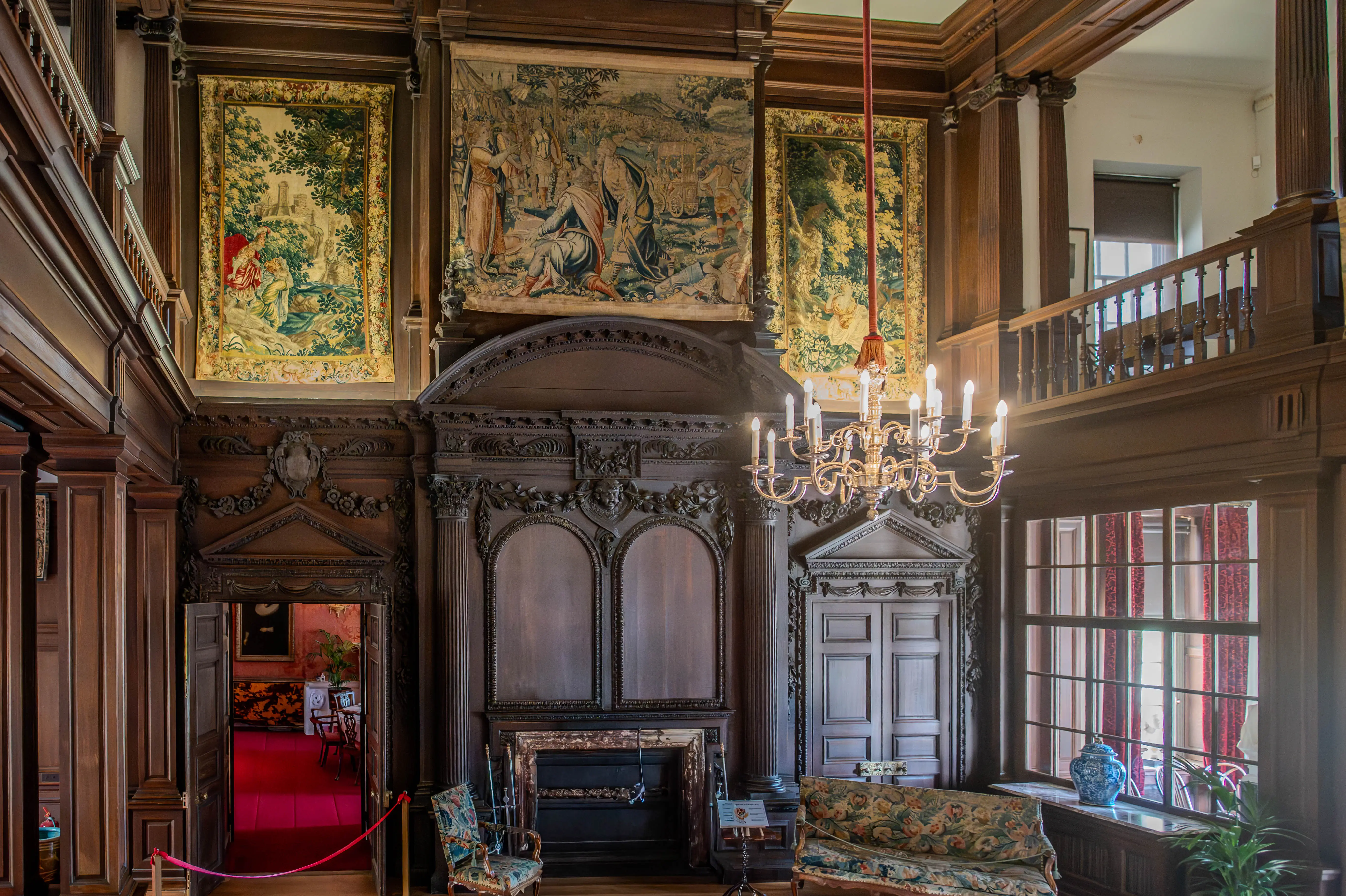 Interior view of a grand hall with three large, detailed tapestries on the wall, a dark wood fireplace and panelling, a multi-armed chandelier, a sofa and chairs with floral patterns, and a balcony overlooking the room.