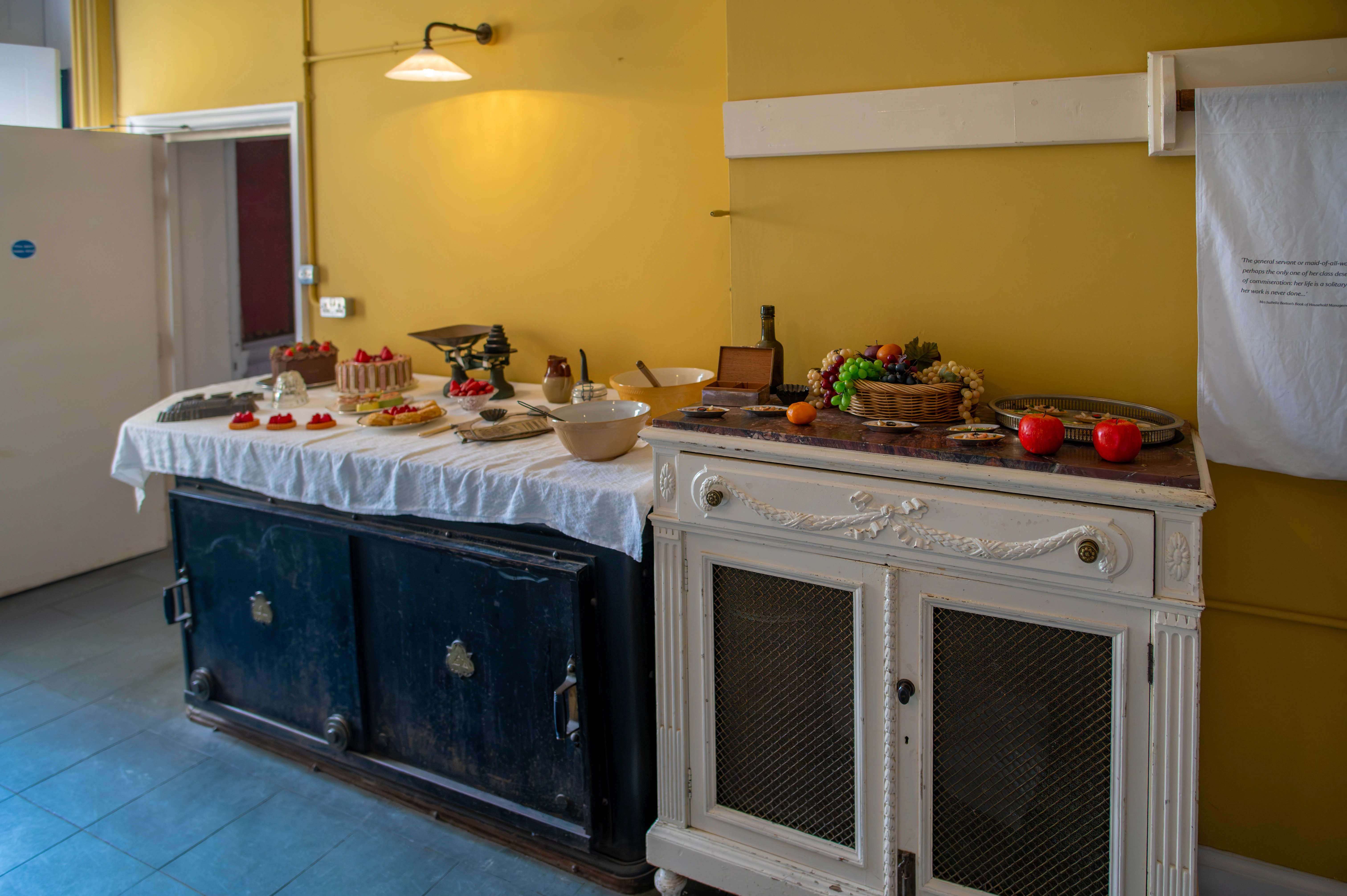 A long kitchen counter covered with a white tablecloth displays several cakes and pastries, alongside bowls, a scale, and a basket of fruit. A vintage cabinet stands to the right, and the walls are painted yellow.
