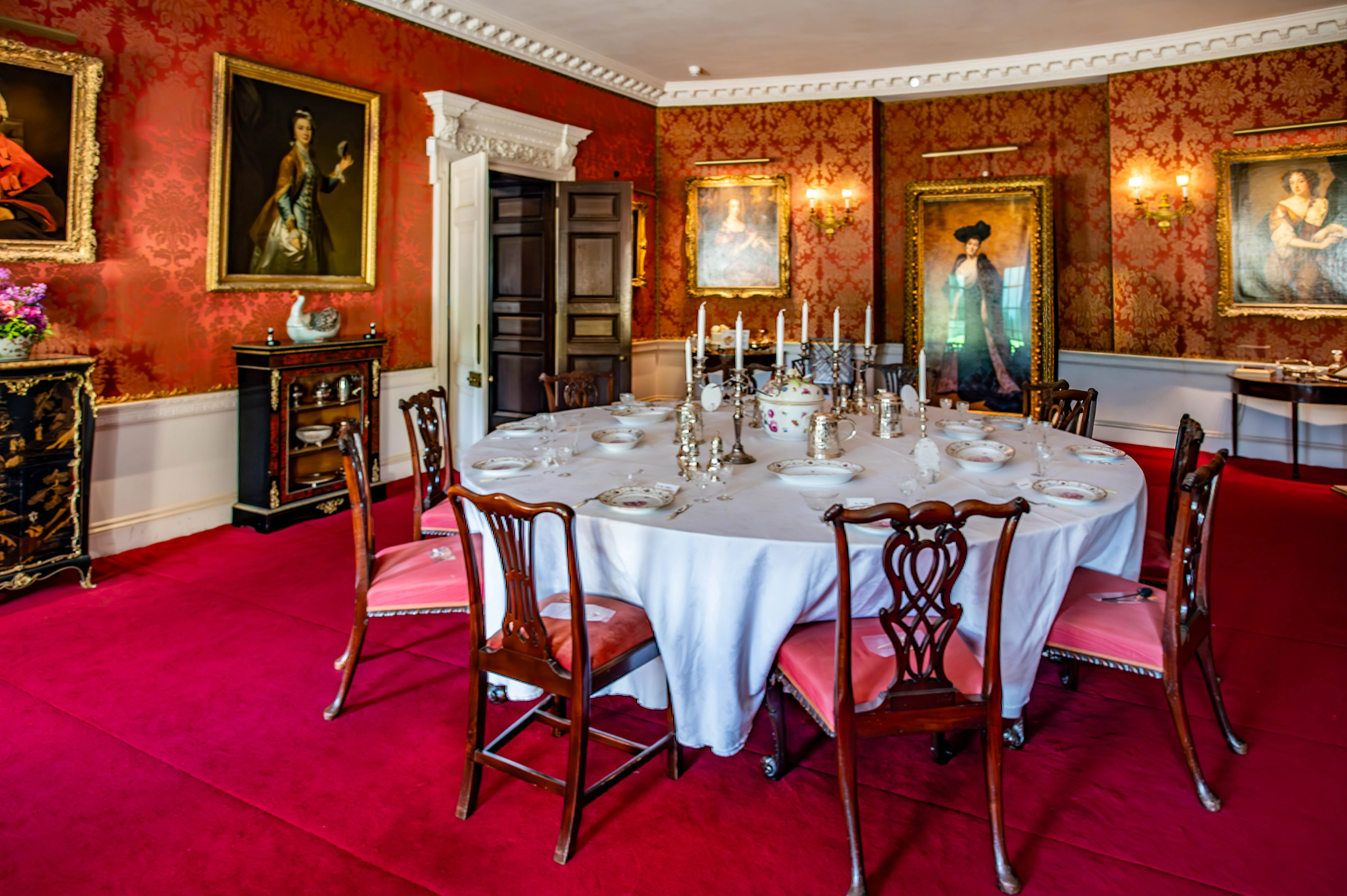 A formal dining room with a large round table set for a meal, surrounded by wooden chairs with pink cushions. The walls are covered in red damask wallpaper and feature several large portraits in gilded frames. The floor is covered in a rich red carpet.