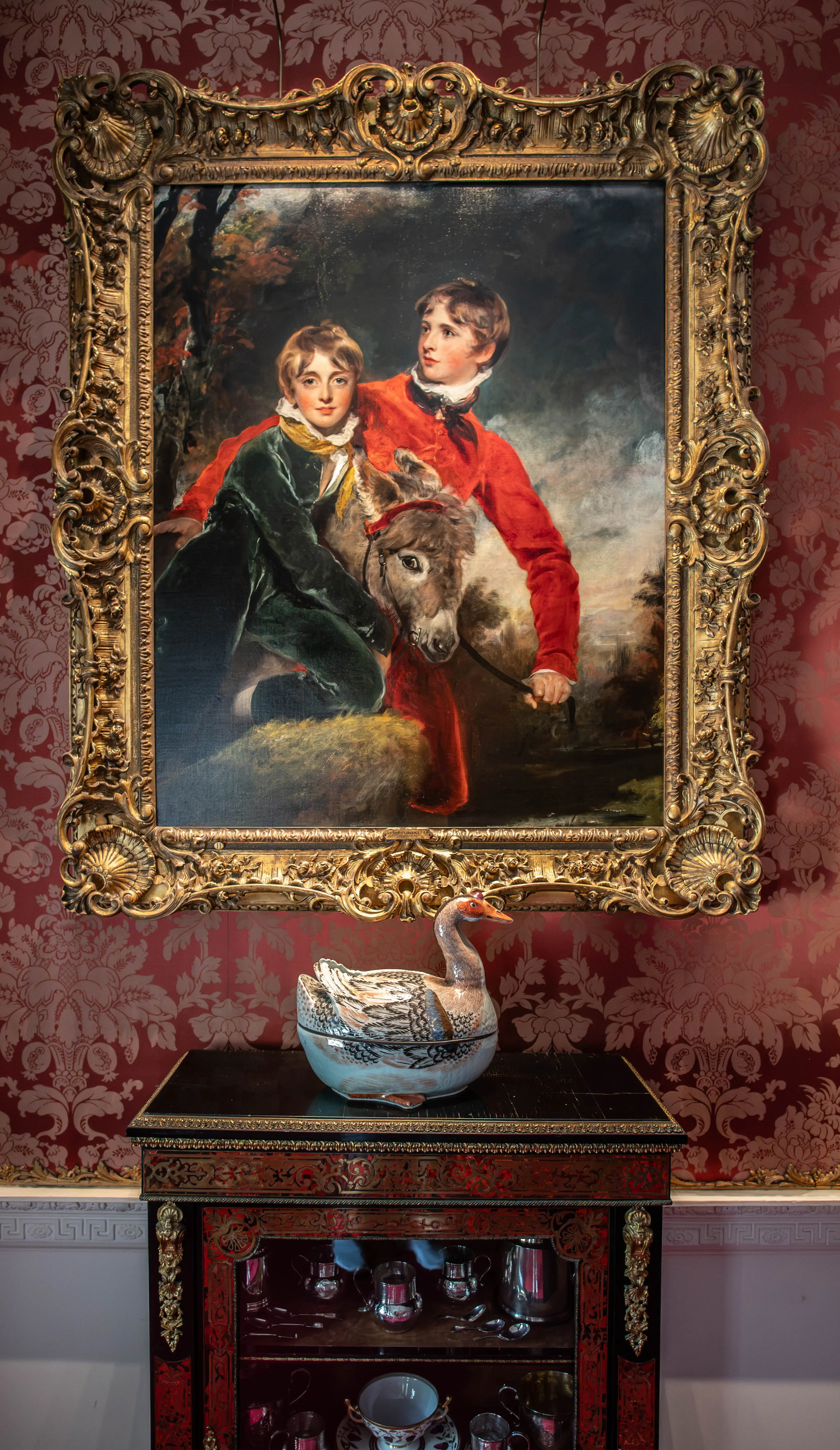 A large, ornate gold-framed painting of two boys and a donkey on a red damask patterned wall, above a black lacquered cabinet with ornate gold and red detailing, containing silver teaware and ornaments. A decorative ceramic swan sits on top of the cabinet.