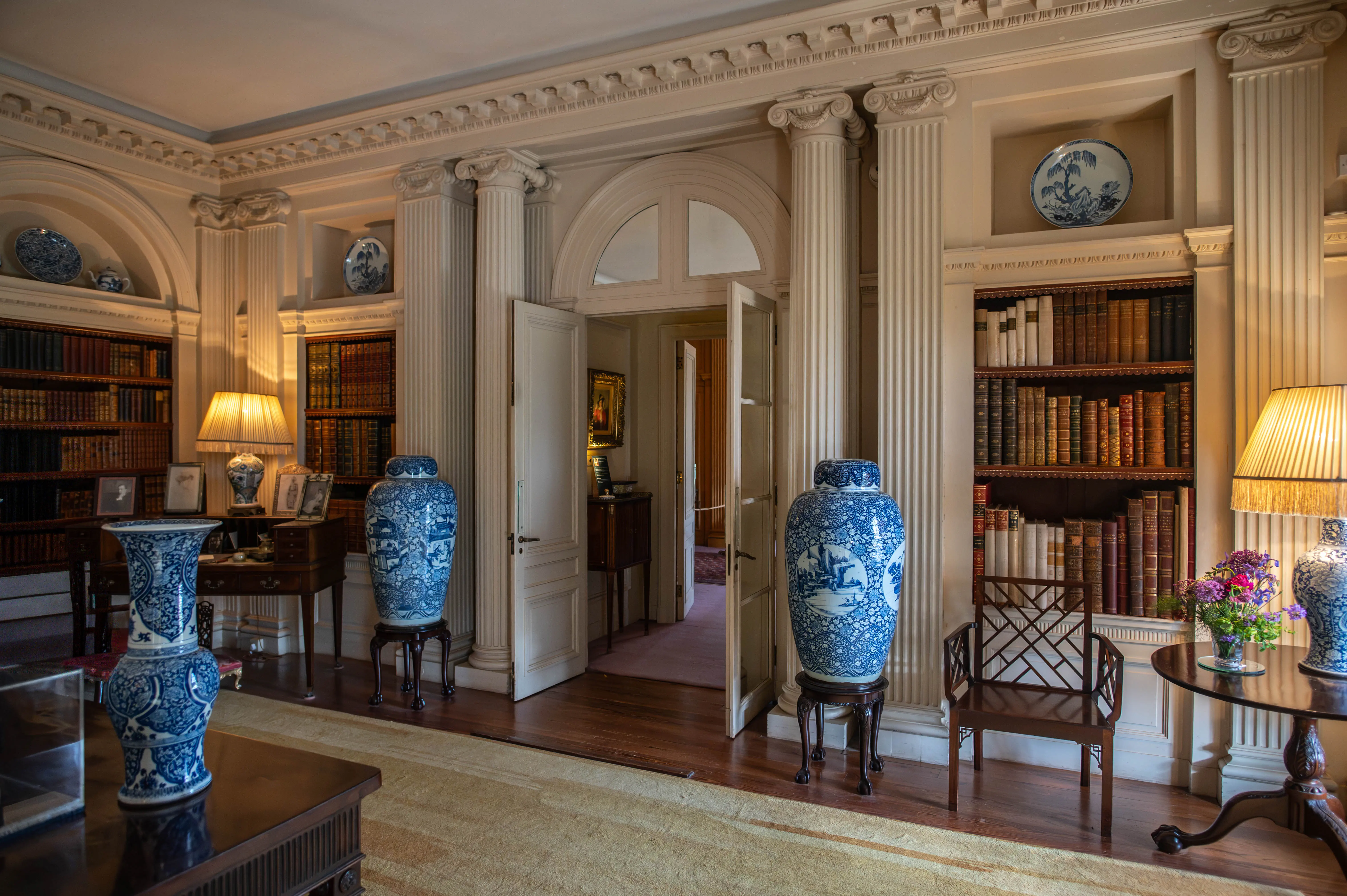 A wide shot of a luxurious library interior. Tall, fluted columns frame bookshelves filled with leather-bound books. Two large blue and white porcelain vases stand on ornate stands. A round side table holds a lamp and a bouquet of flowers.
