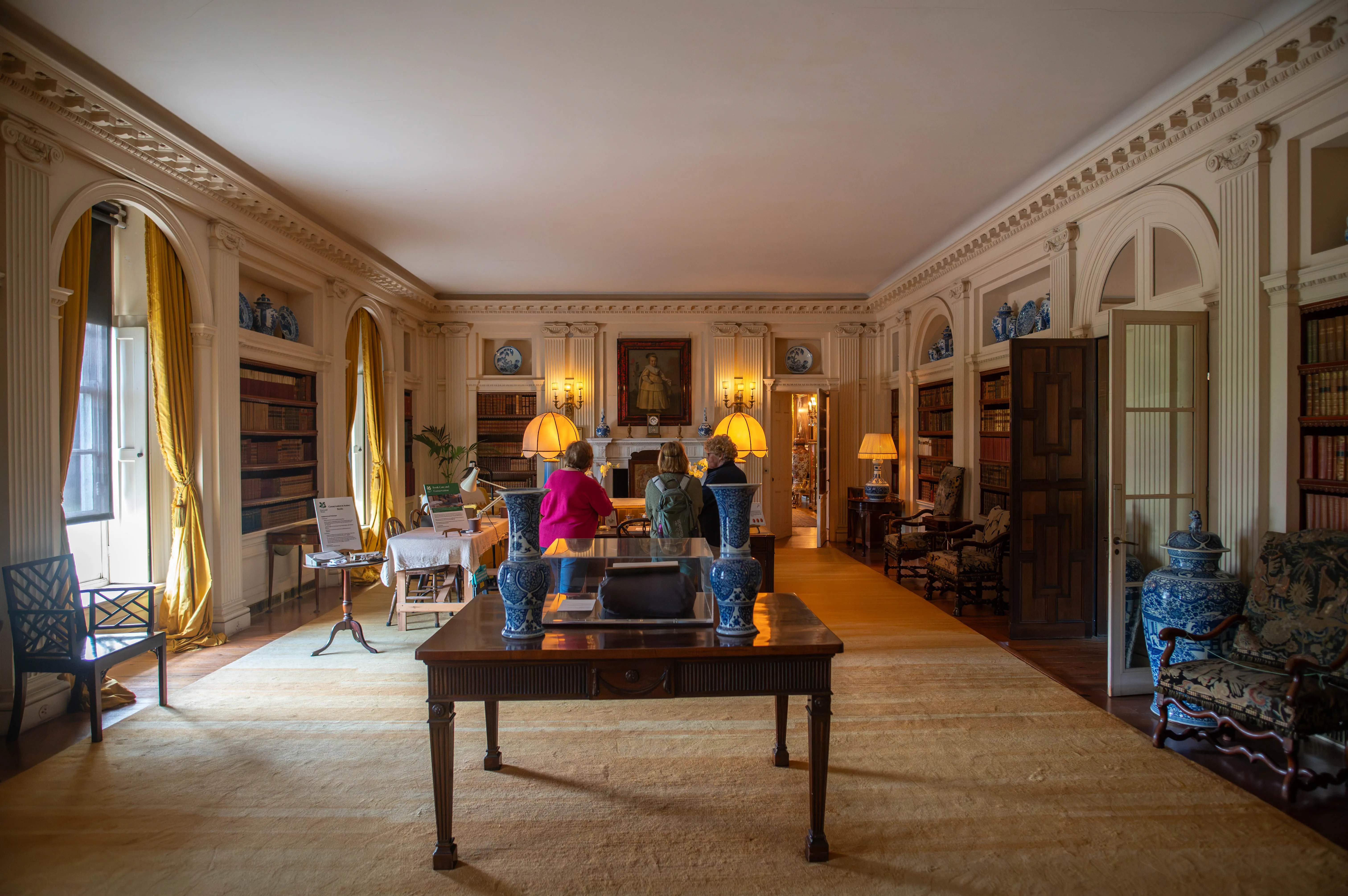 A wide shot of a long, opulent library room with three women standing near a central table. Bookshelves line the walls, and sunlight streams through tall windows.
