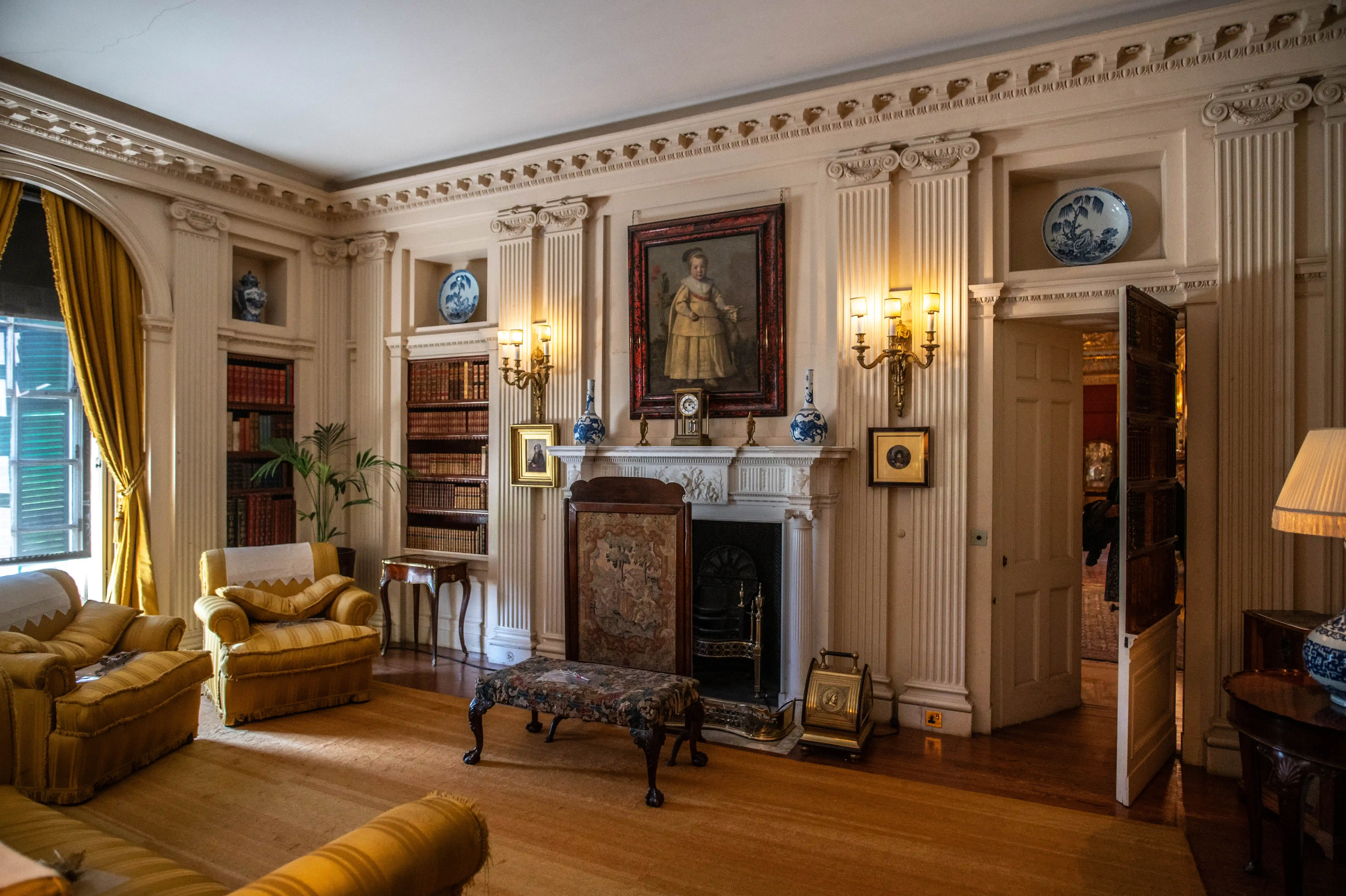 A view of a drawing room with yellow upholstered furniture, two bookcases filled with books, a fireplace with a portrait above it, and decorative vases.