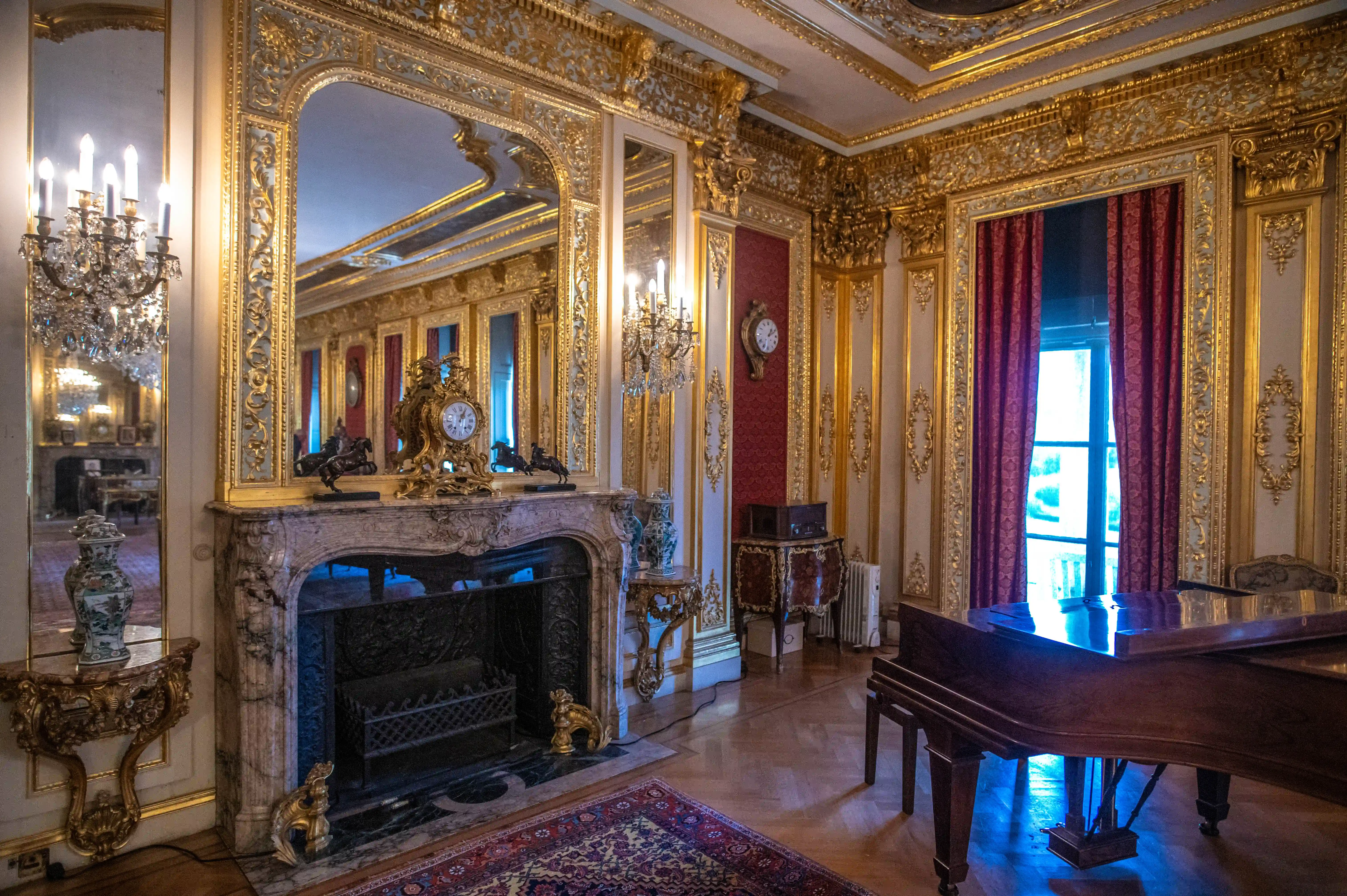 A well-lit, ornate drawing-room interior showcasing a large marble fireplace with a gold clock and decorative elements, a dark wood grand piano reflecting blue light, and gilded walls with deep red drapery covering a tall window.