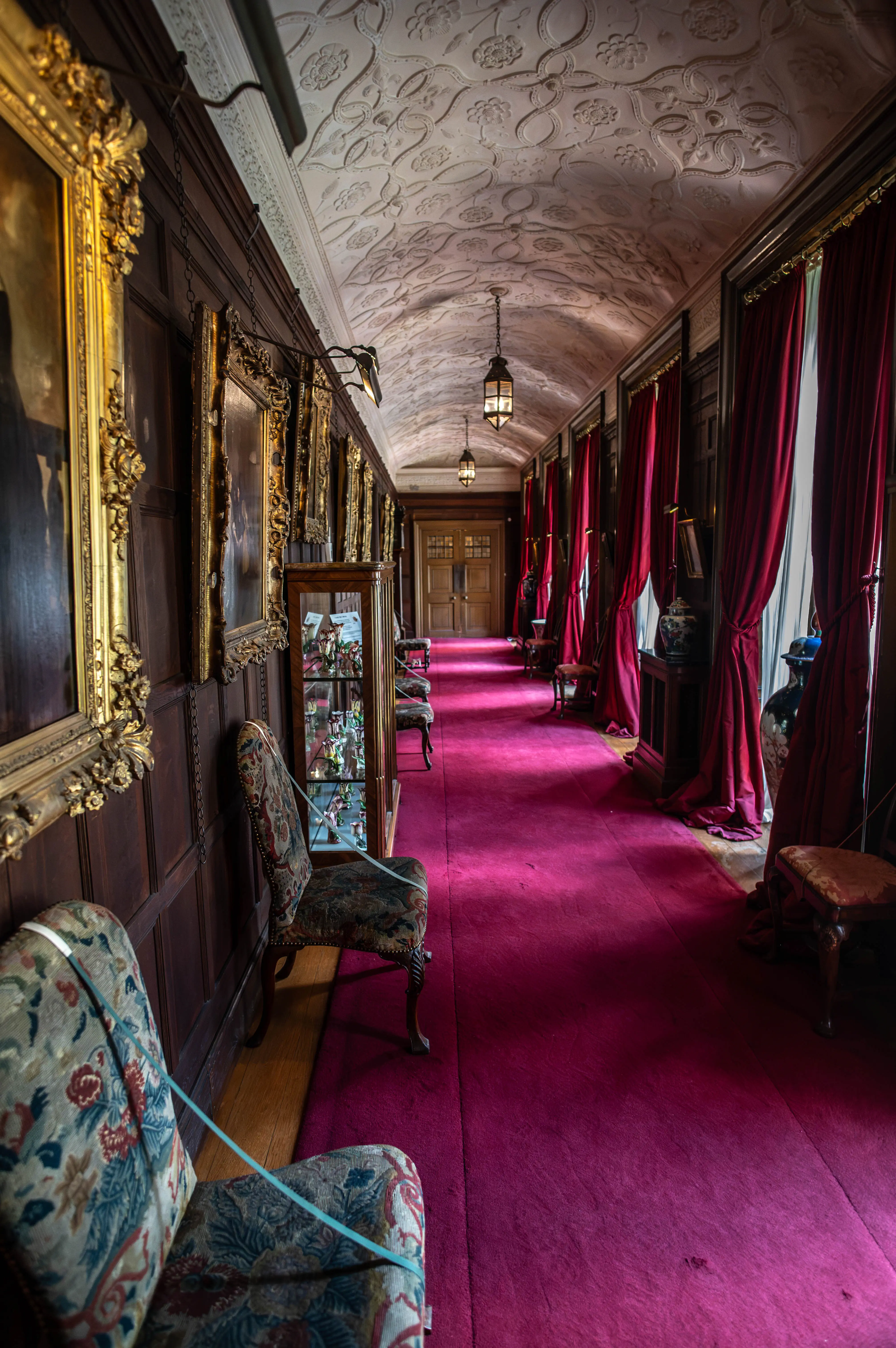 A long, wide hallway in a stately home, with a rich magenta carpet, dark wood panelling, and plush red curtains. Gold-framed paintings hang on the walls and a decorative patterned ceiling is visible.