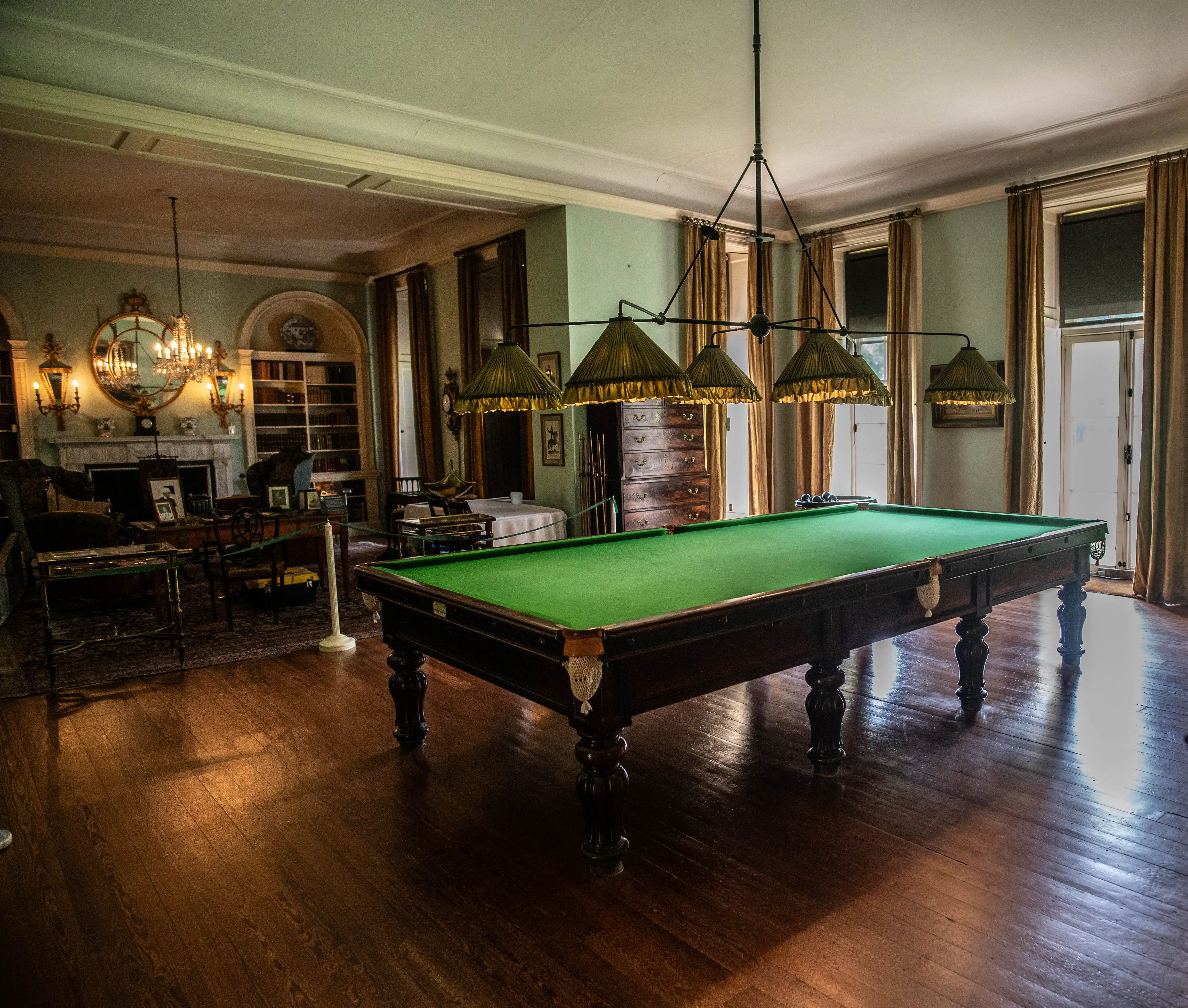 A dimly lit, luxurious billiard room with a green snooker table, chandeliers, and antique furniture.