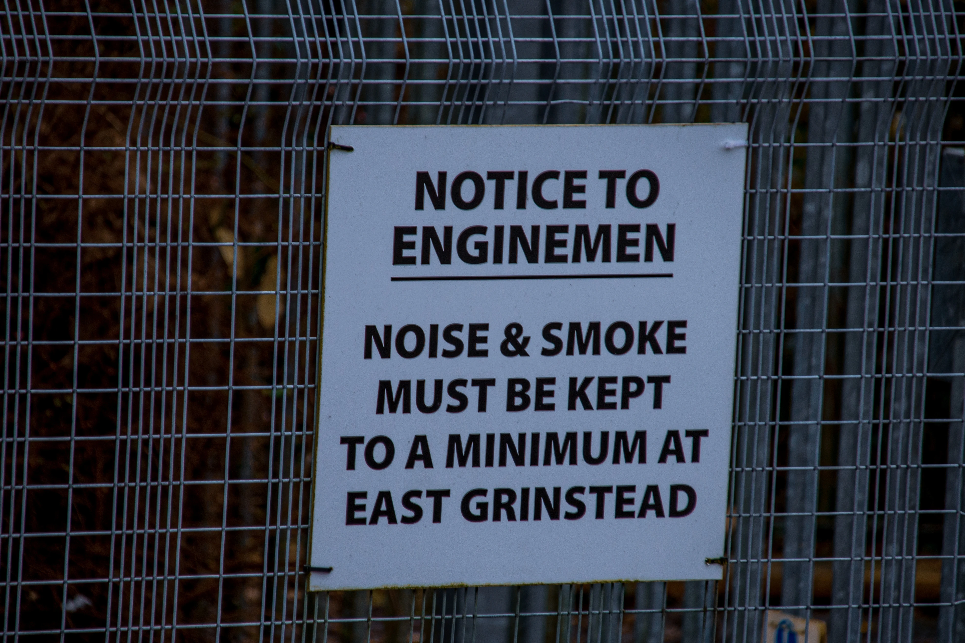 A white sign with bold black text attached to a metal wire fence. The sign reads: NOTICE TO ENGINEMEN. NOISE & SMOKE MUST BE KEPT TO A MINIMUM AT EAST GRINSTEAD.
