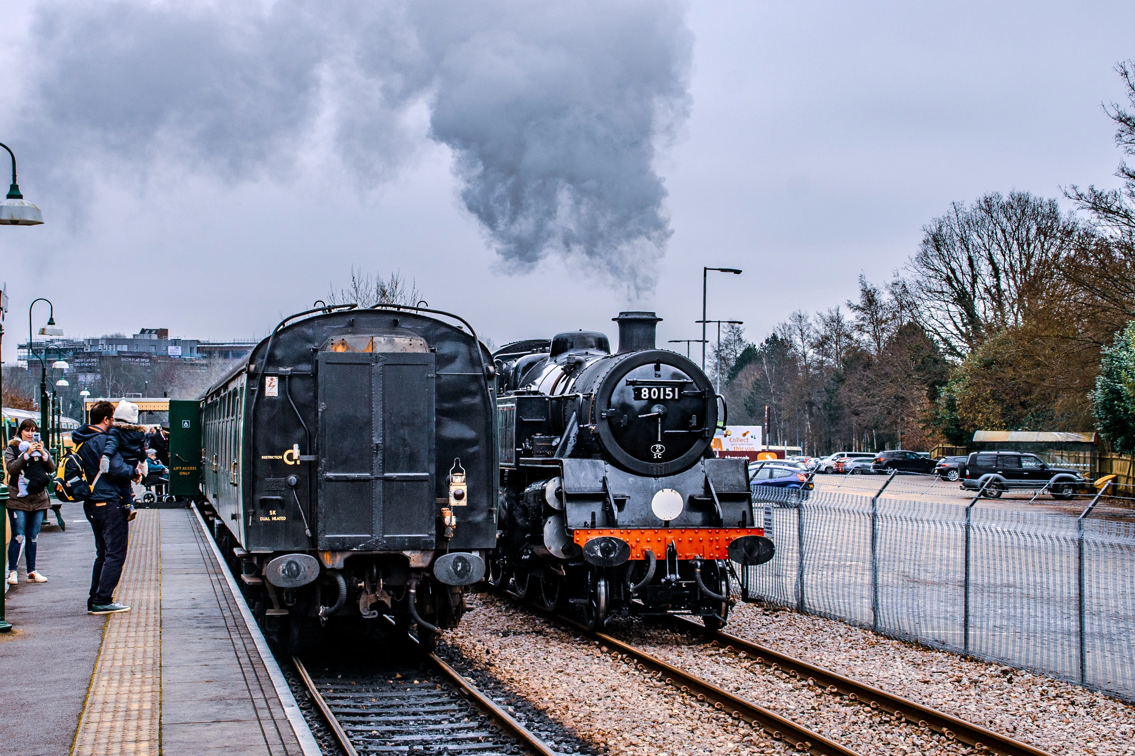 A black steam locomotive with red detailing is pulling a train of old-fashioned carriages along a railway track. Dark smoke is billowing from the chimney. The train is at a station platform with green benches and flowerbeds.