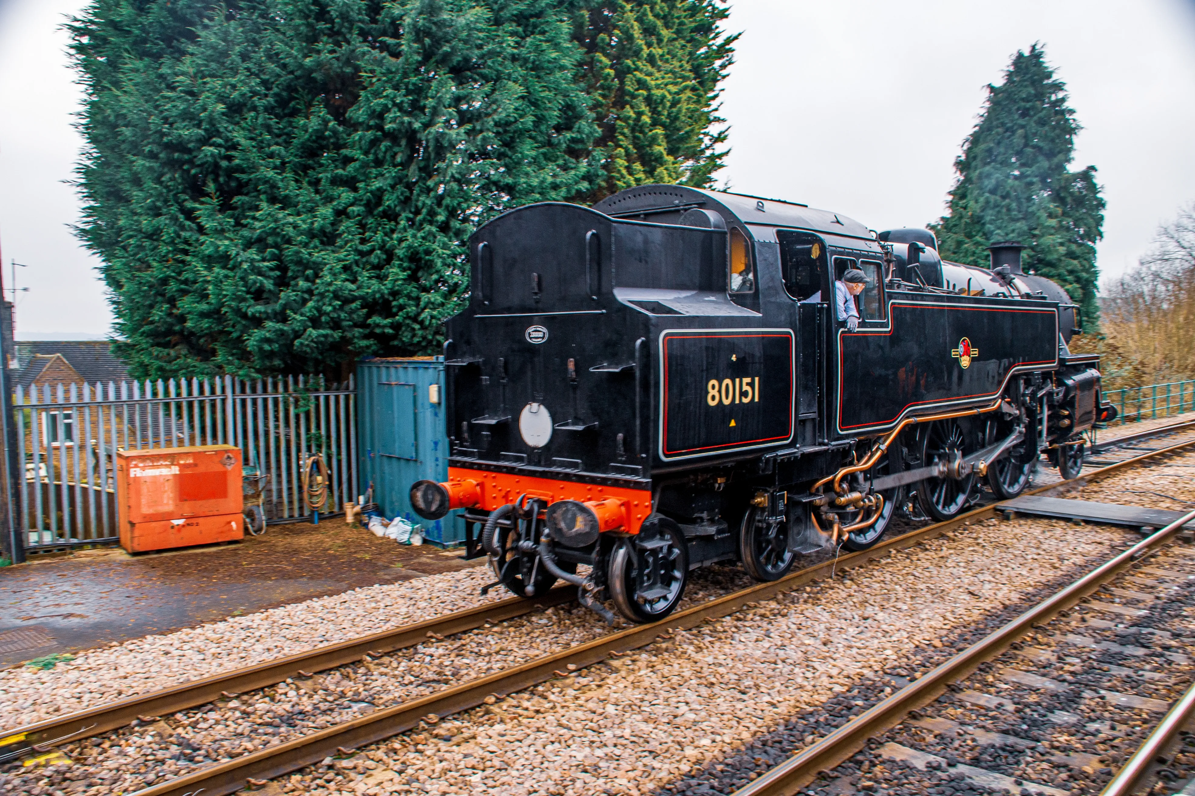 A vintage steam locomotive, No. 80151, stationed on railway tracks. The locomotive has a black body with red and gold accents, a red undercarriage, and intricate details like pipes, wheels, and a chimney. A person is visible in the cabin. The background includes a metal fence, trees, an orange toolbox, and a blue container