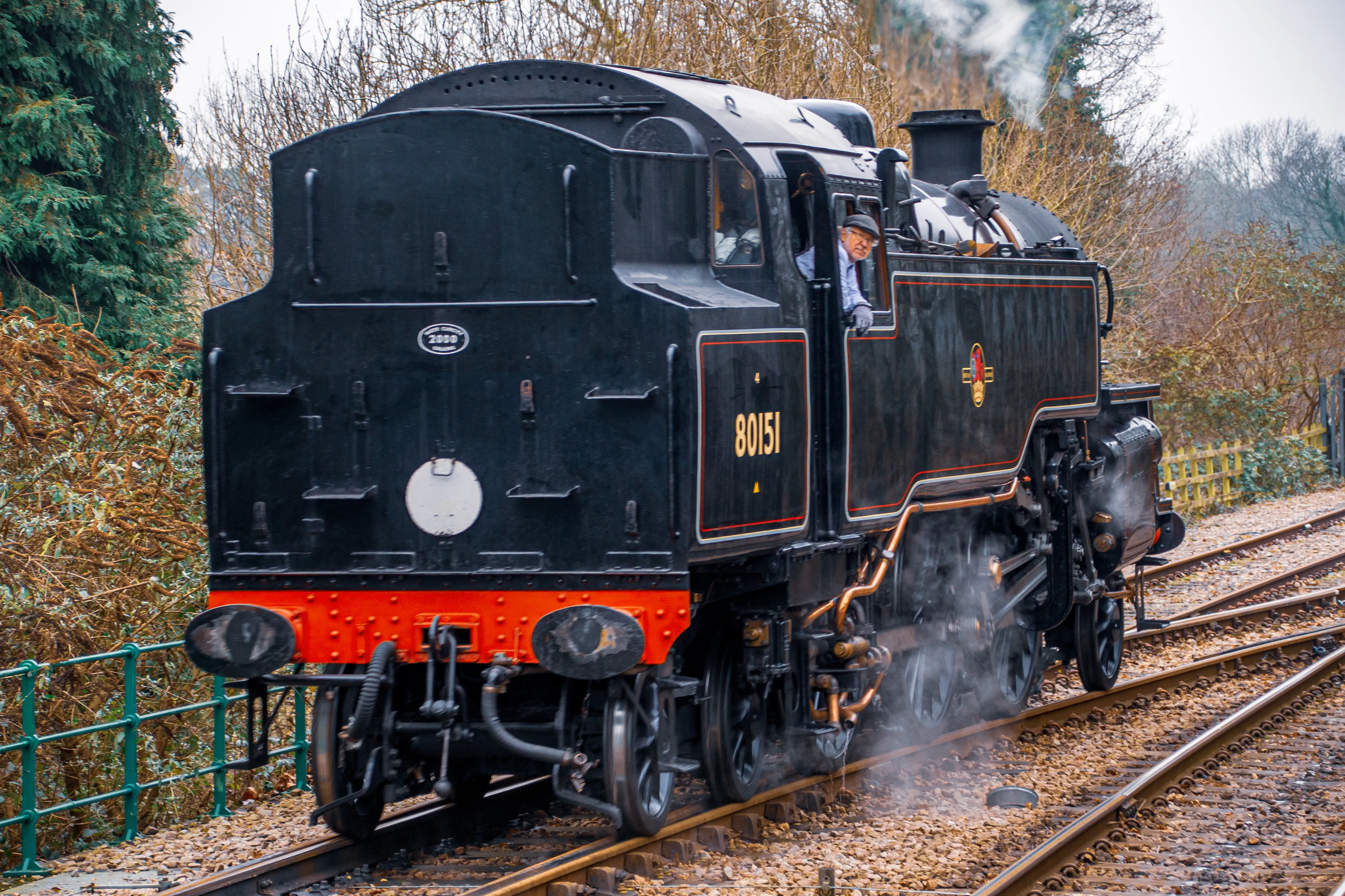 A black steam locomotive with the number 80151 on its side is moving on a railway track. A driver in a cap is looking out of the cab window and smiling. Steam is coming from the engine.
