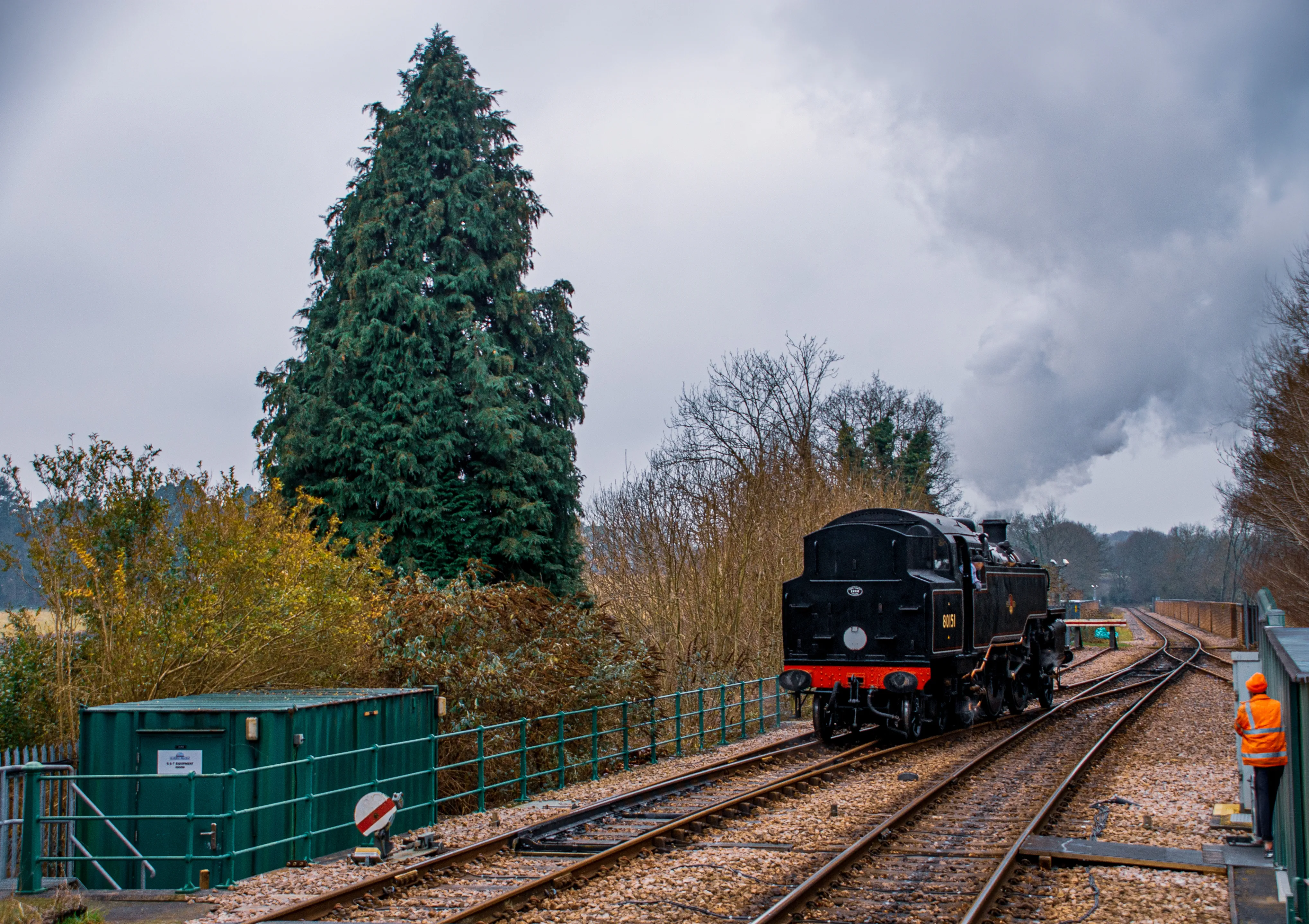 A black steam train numbered 80151 is moving along tracks, emitting steam, with a railway worker in an orange jacket visible on the right.