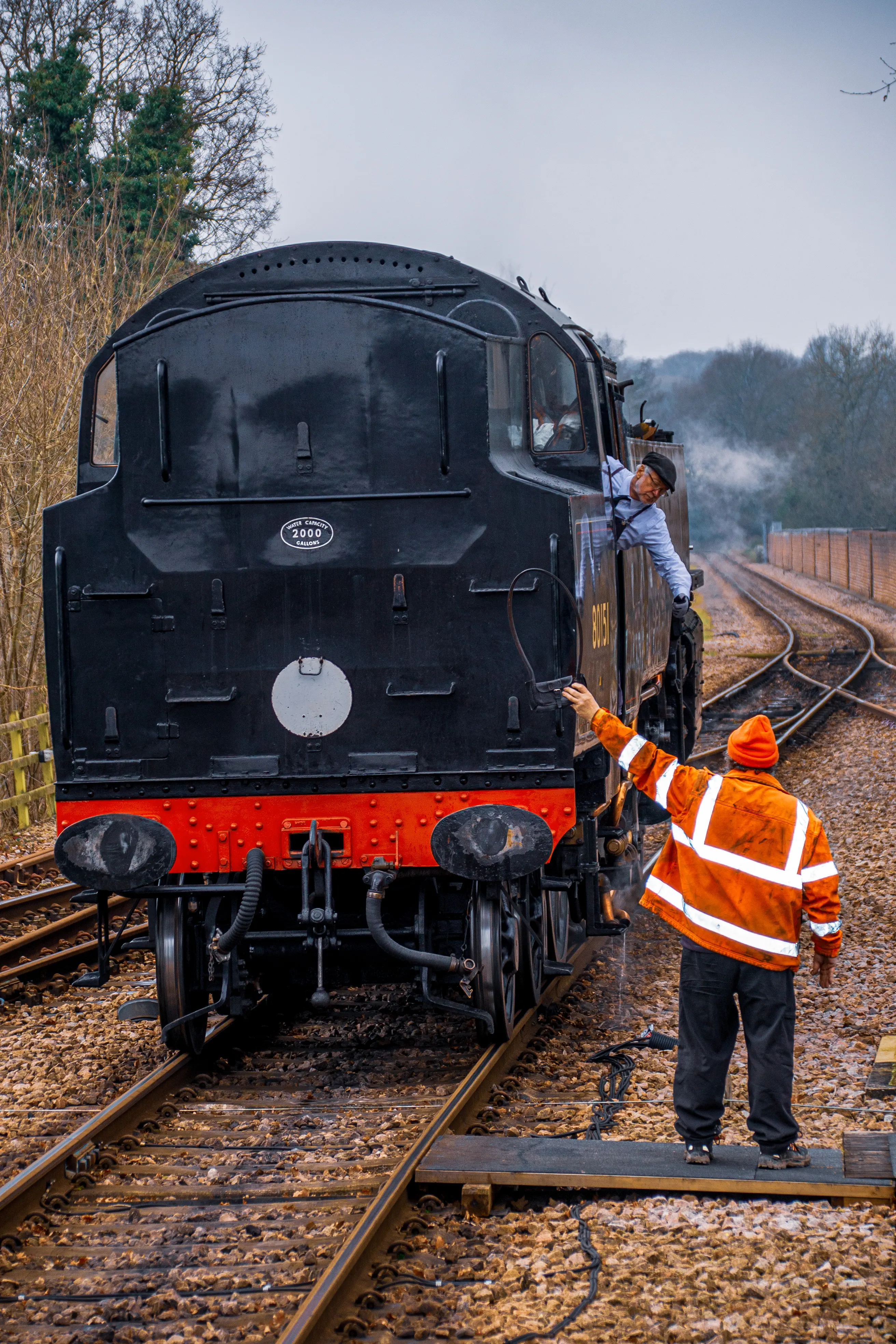 A railway worker in an orange high-visibility jacket and beanie signals towards a steam train driver who is leaning out of the cab window. The steam train is black with a red buffer beam and is on a railway track.
